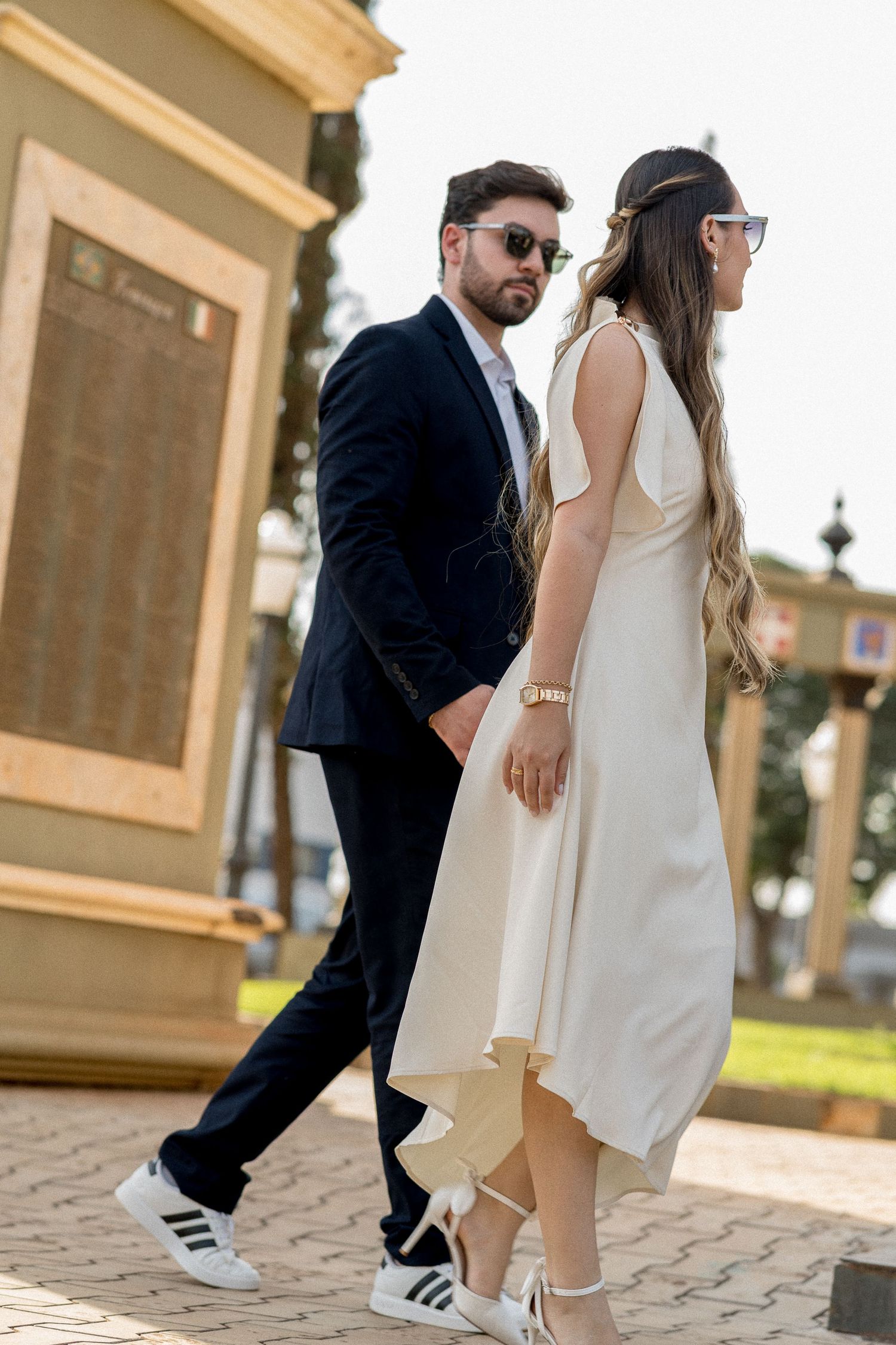 Elegant couple walking outdoors, man in navy suit with sneakers, woman in white midi dress with heels and sunglasses.