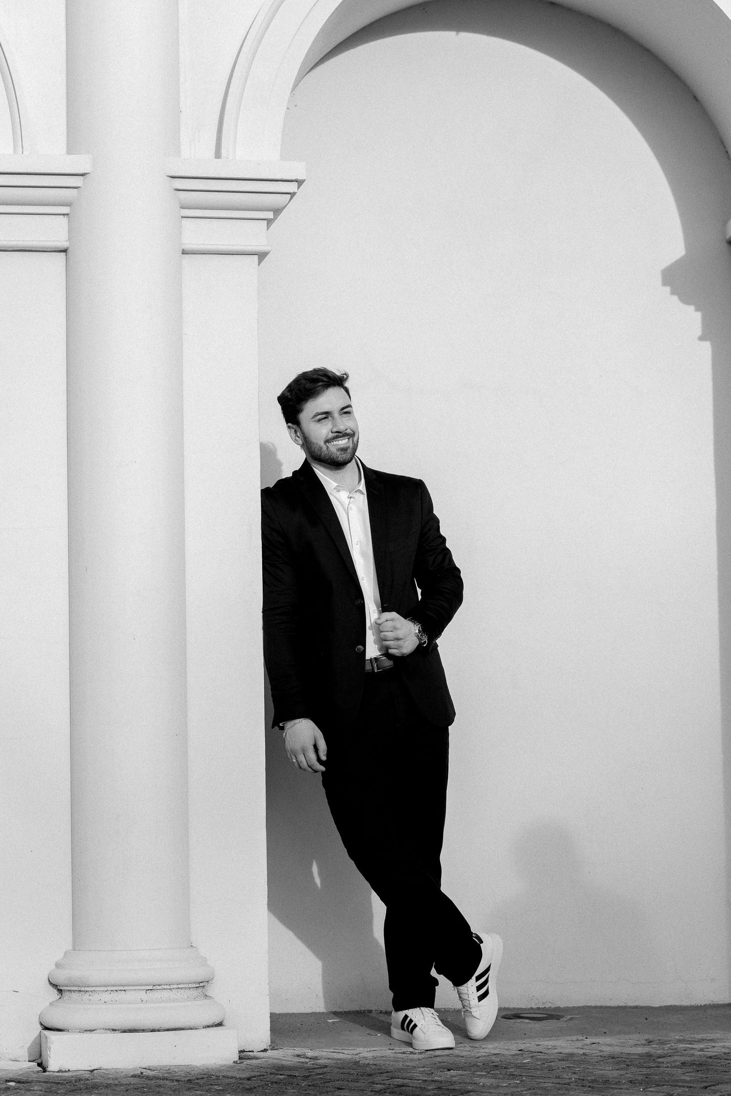 Black and white photo of a smiling man in a suit leaning against a white pillar under an arch.
