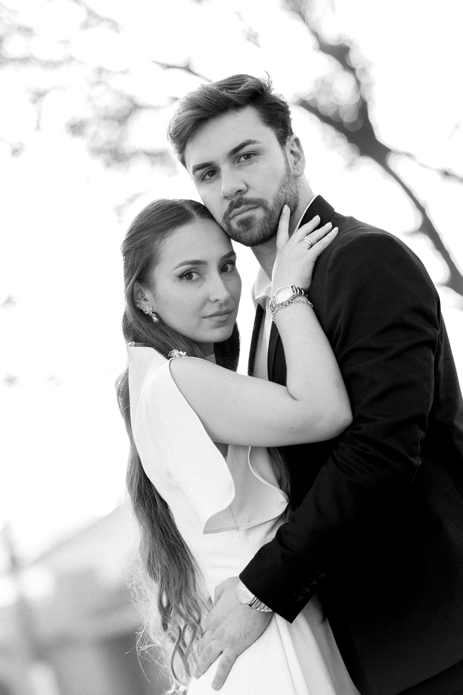 Black and white photo of an elegant couple embracing outdoors, woman in white dress and man in dark suit, romantic wedding portrait.