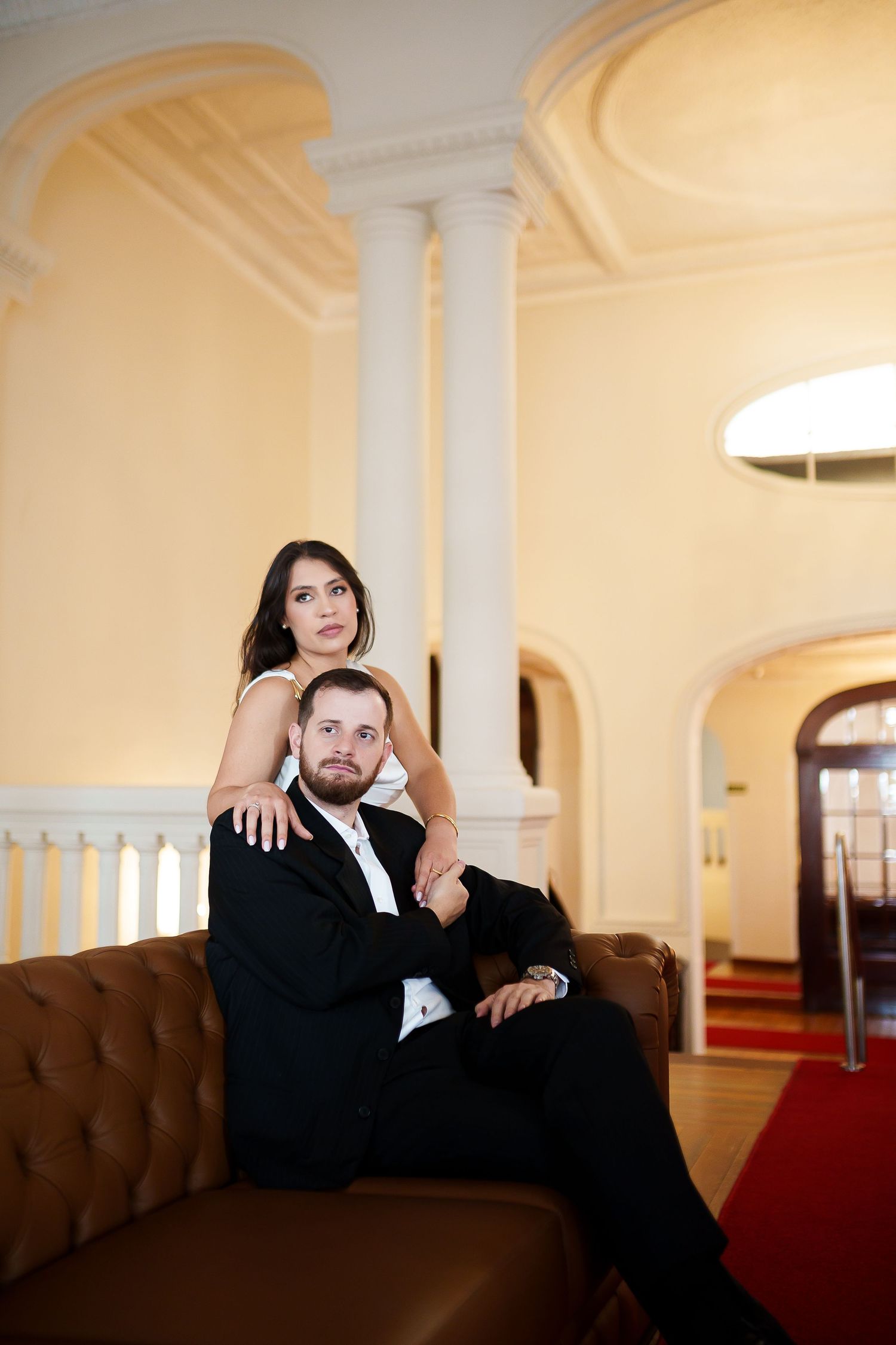 Elegant couple in formal attire posing on a leather sofa in a grand hall with white columns and arched doorways.