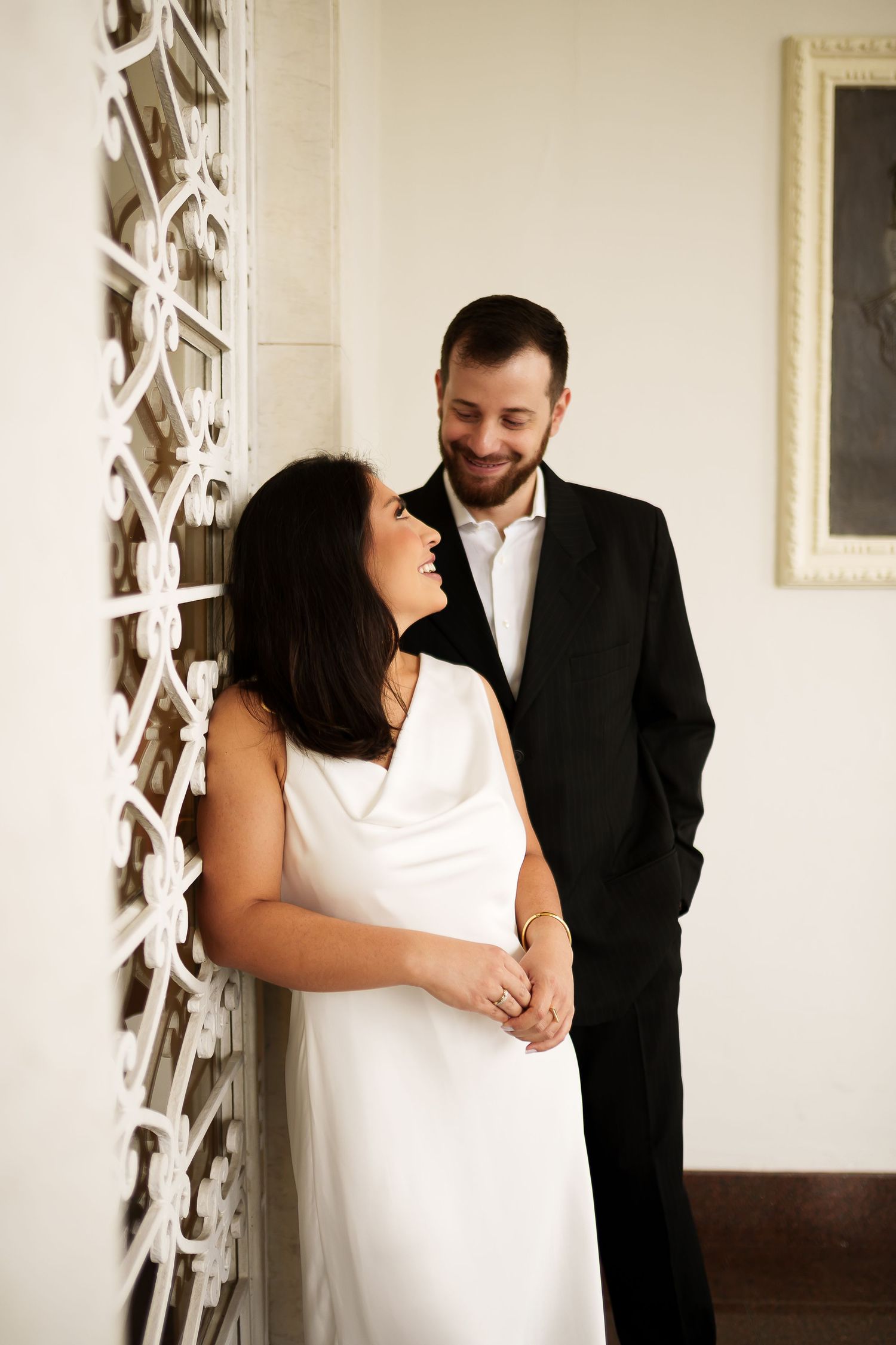 Couple in wedding attire smiling at each other, woman in white dress, man in black suit, leaning against ornate white wall.