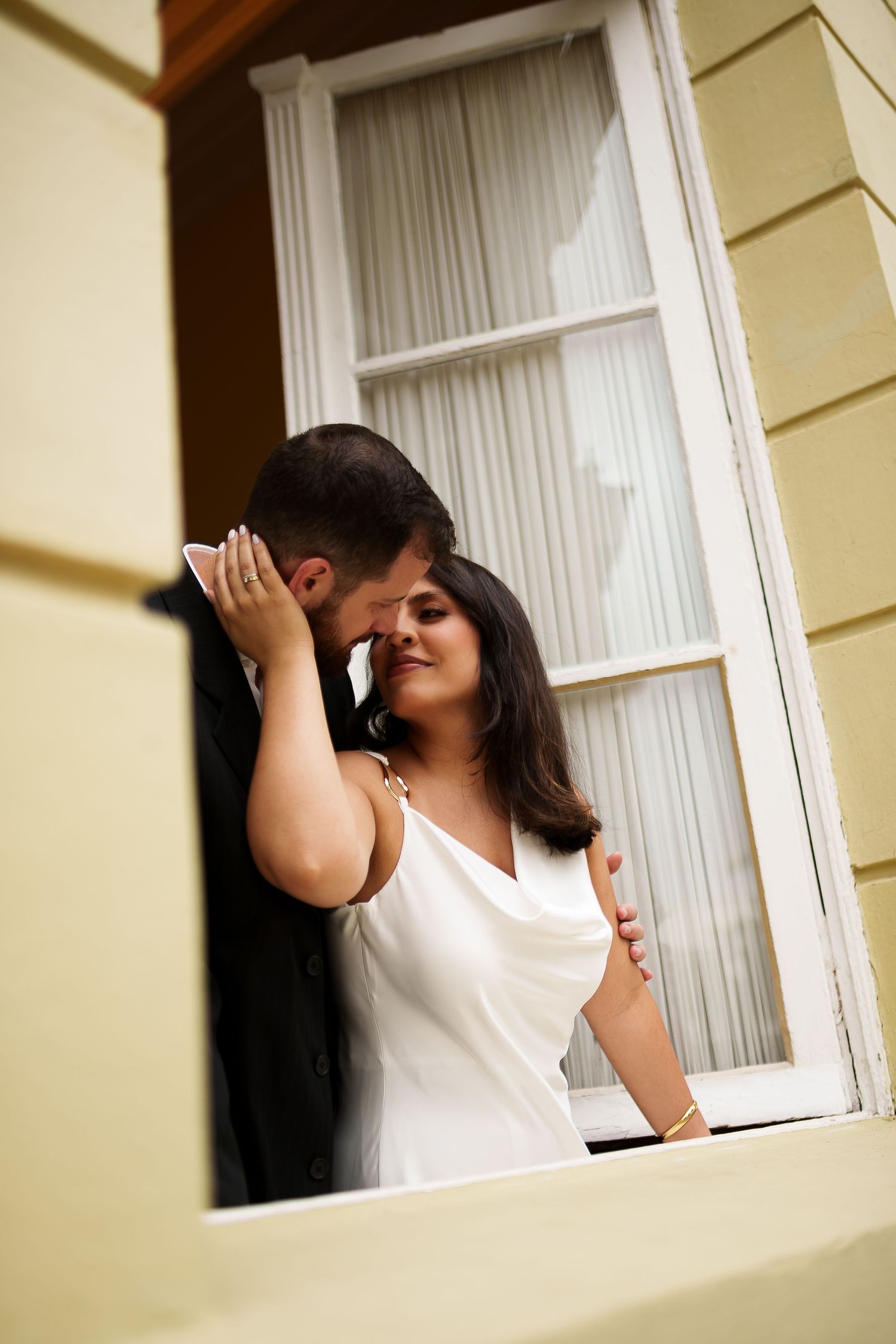 Romantic couple sharing an intimate moment in a yellow building window, woman in white dress with man in dark suit.
