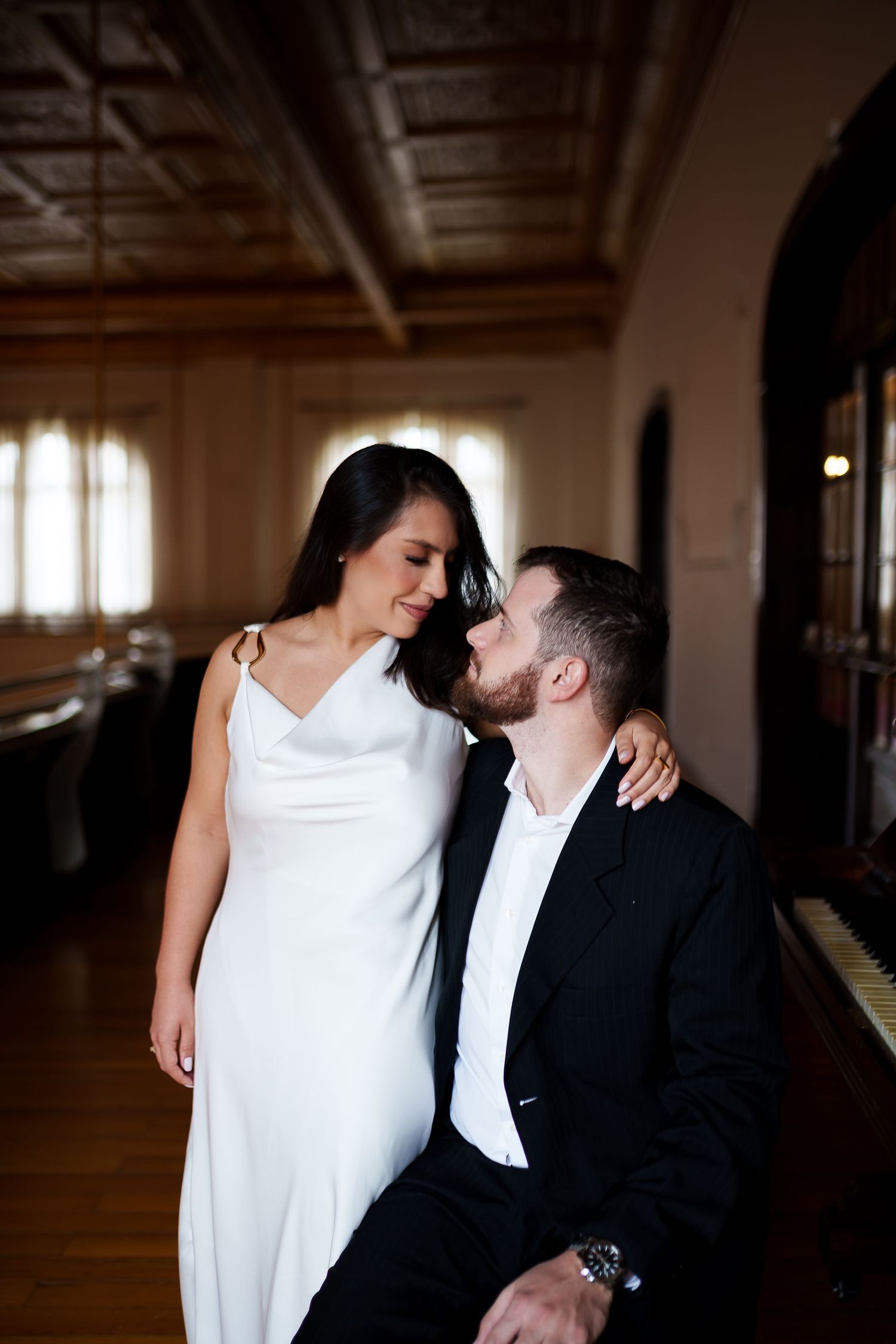 A couple in formal attire share a romantic moment, woman in white dress and man in black suit, in an elegant room.