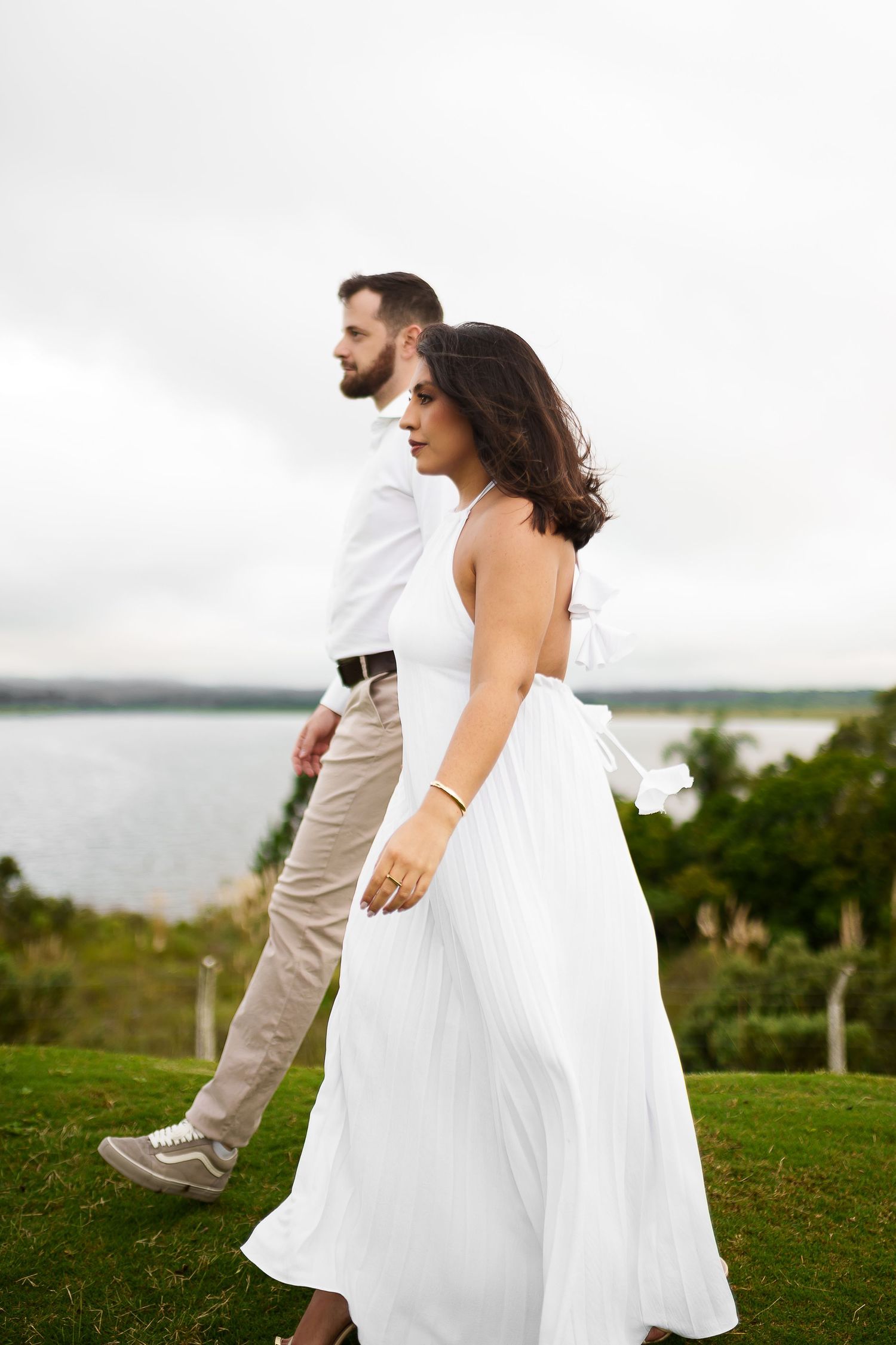 Couple in white outfits walking on green grass near a lake, woman in flowing white dress glances back.