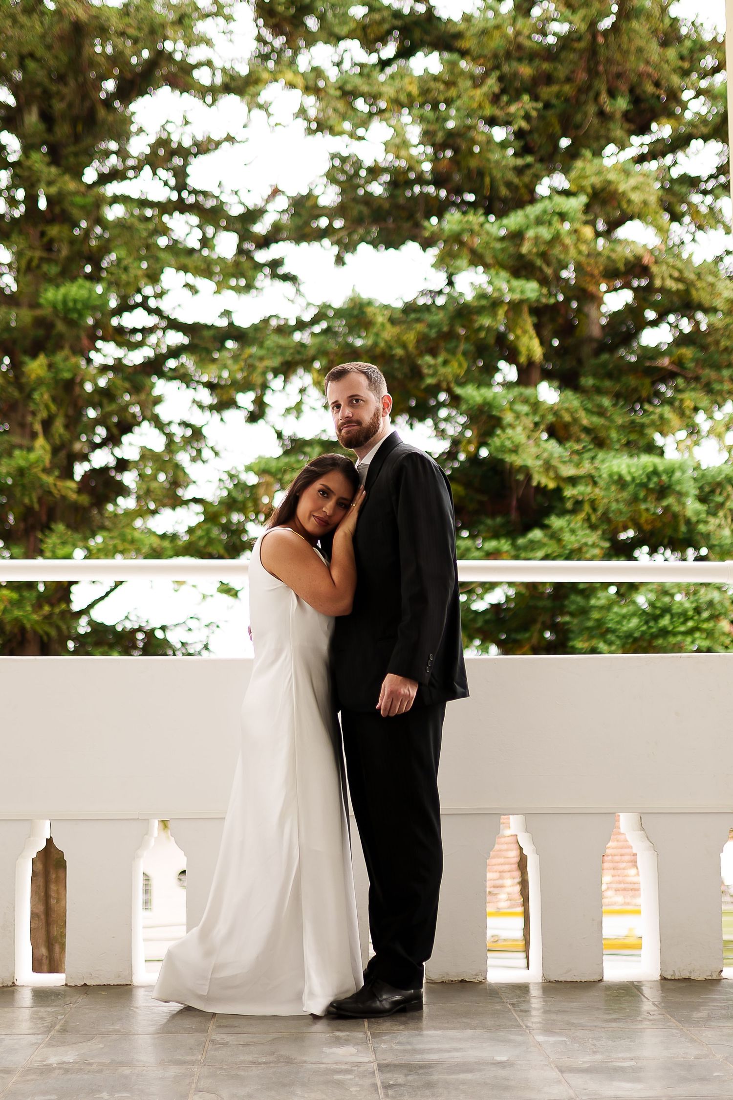 Bride in white gown leans on groom in black suit on elegant white balcony with lush green trees behind them.