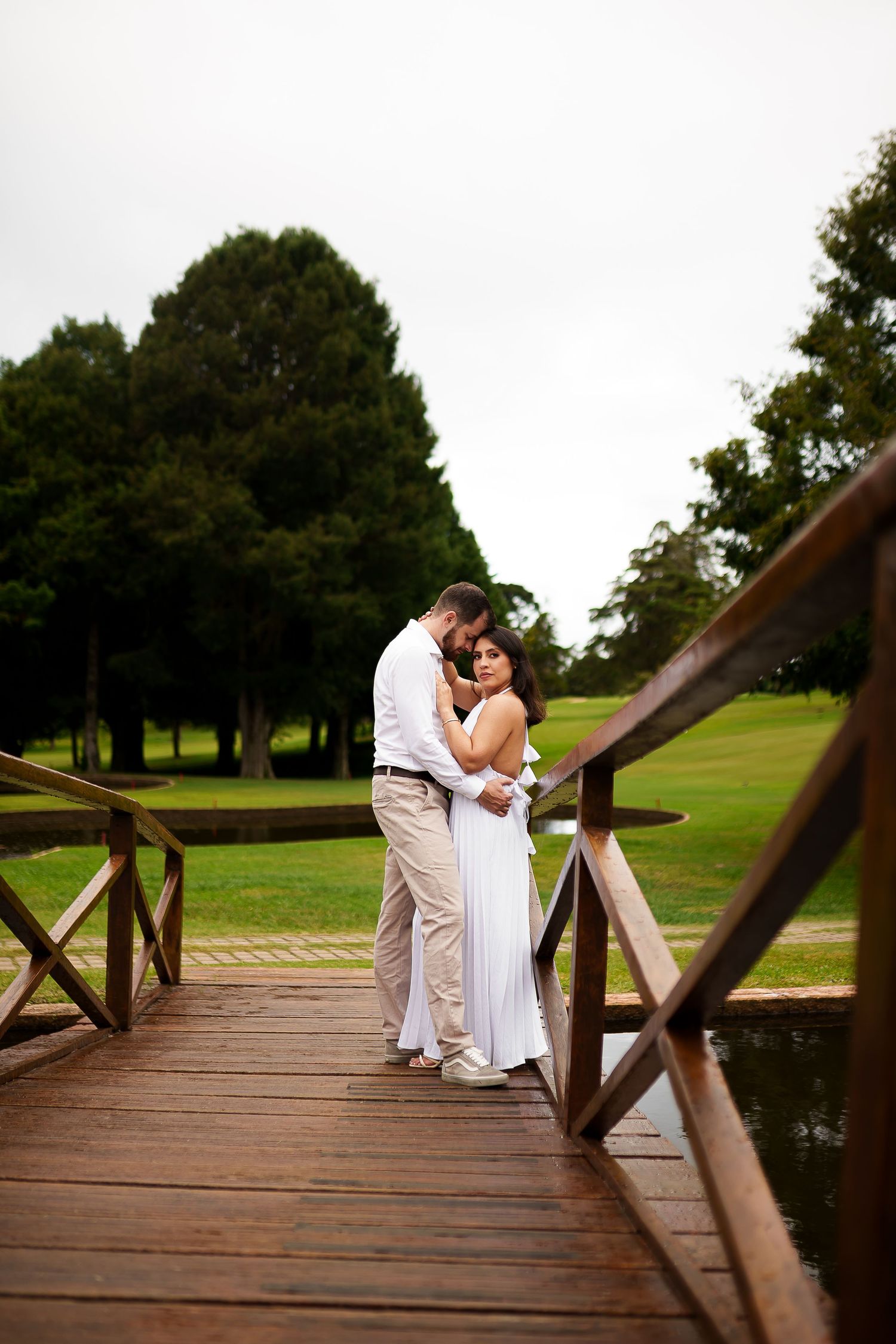 Couple embracing on a wooden bridge surrounded by lush green trees and grass on a cloudy day.