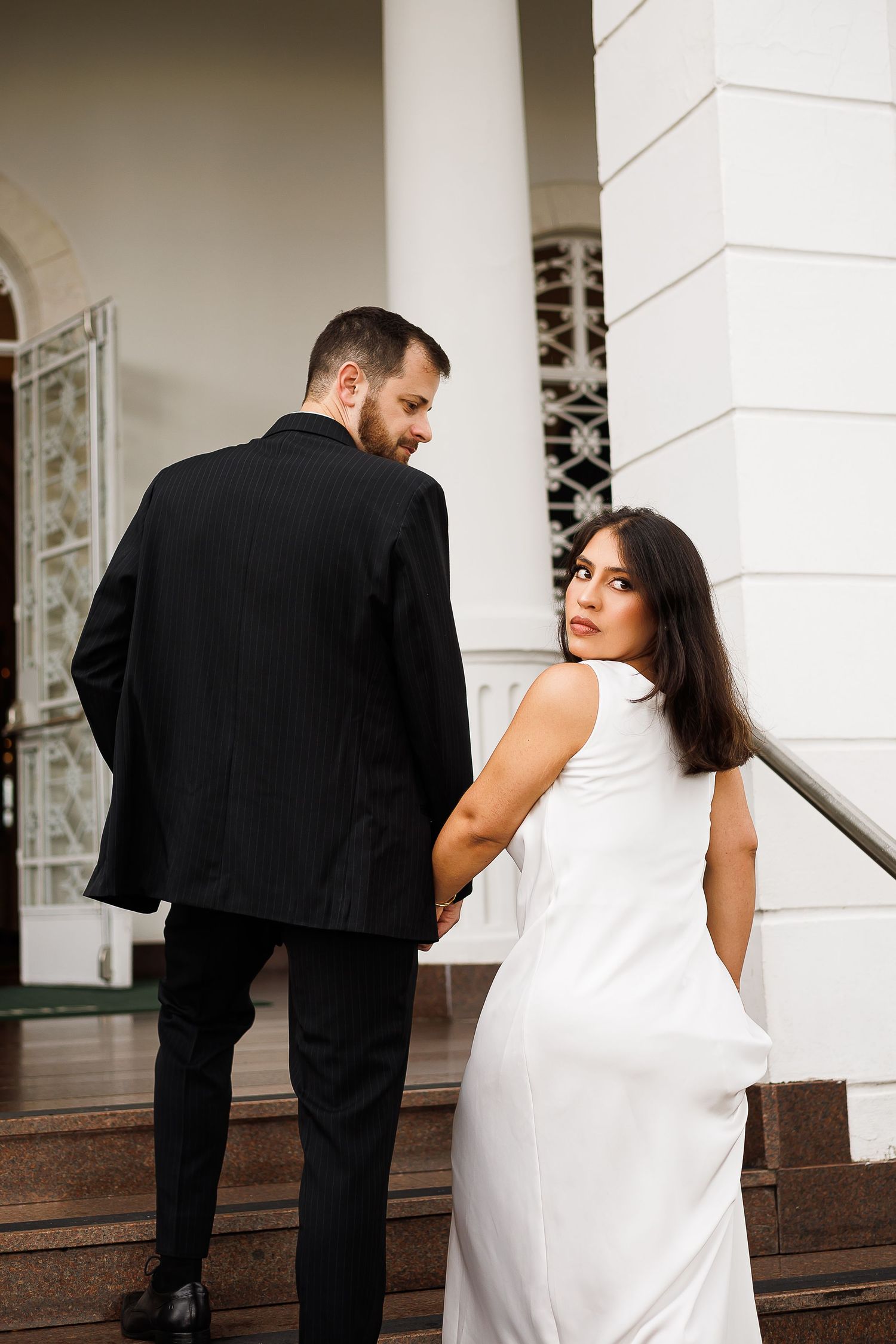 Elegant couple in black suit and white dress holding hands on steps of a white building.