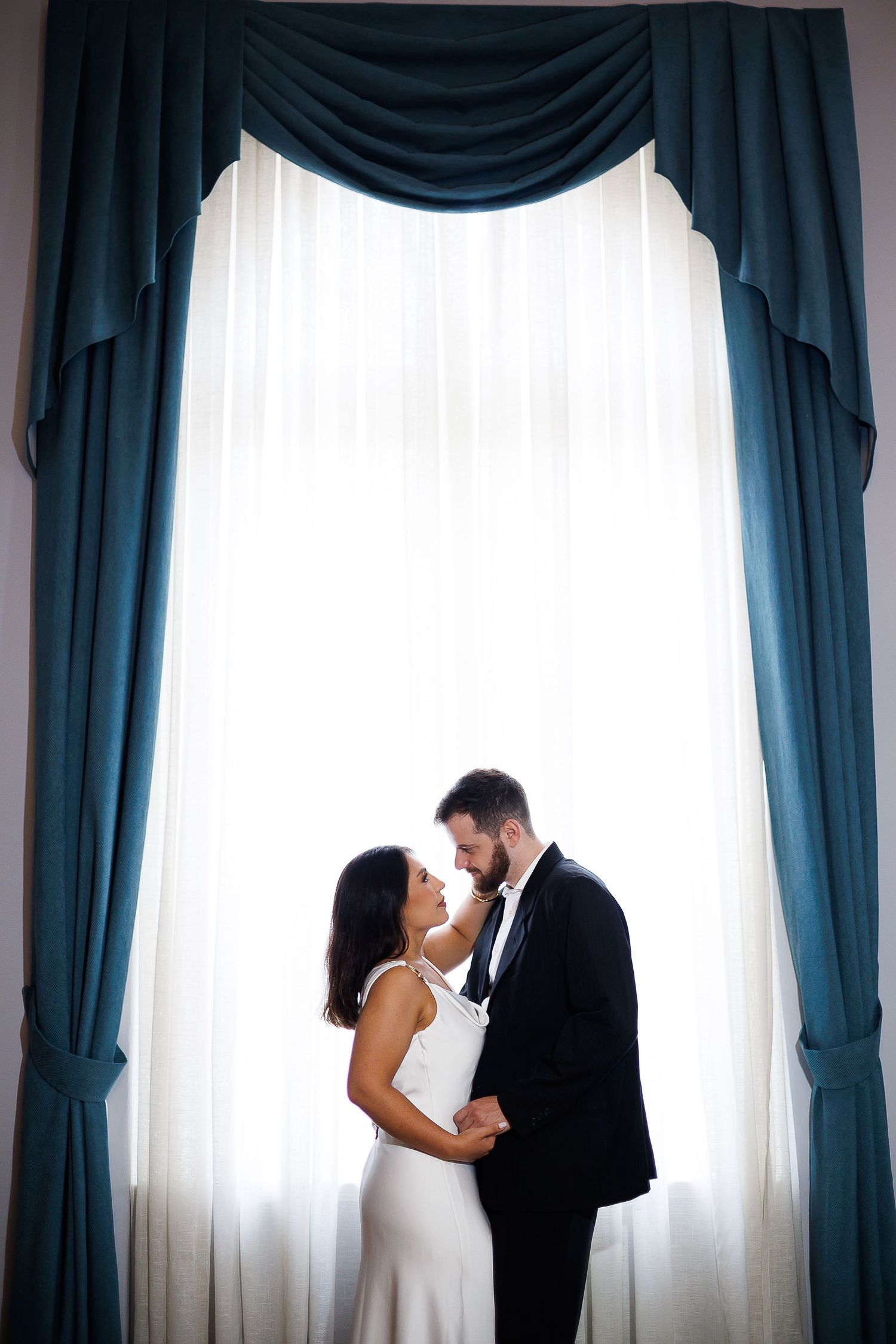 Elegant wedding couple standing intimately before a grand window with teal drapes, bride in white gown, groom in black tuxedo.