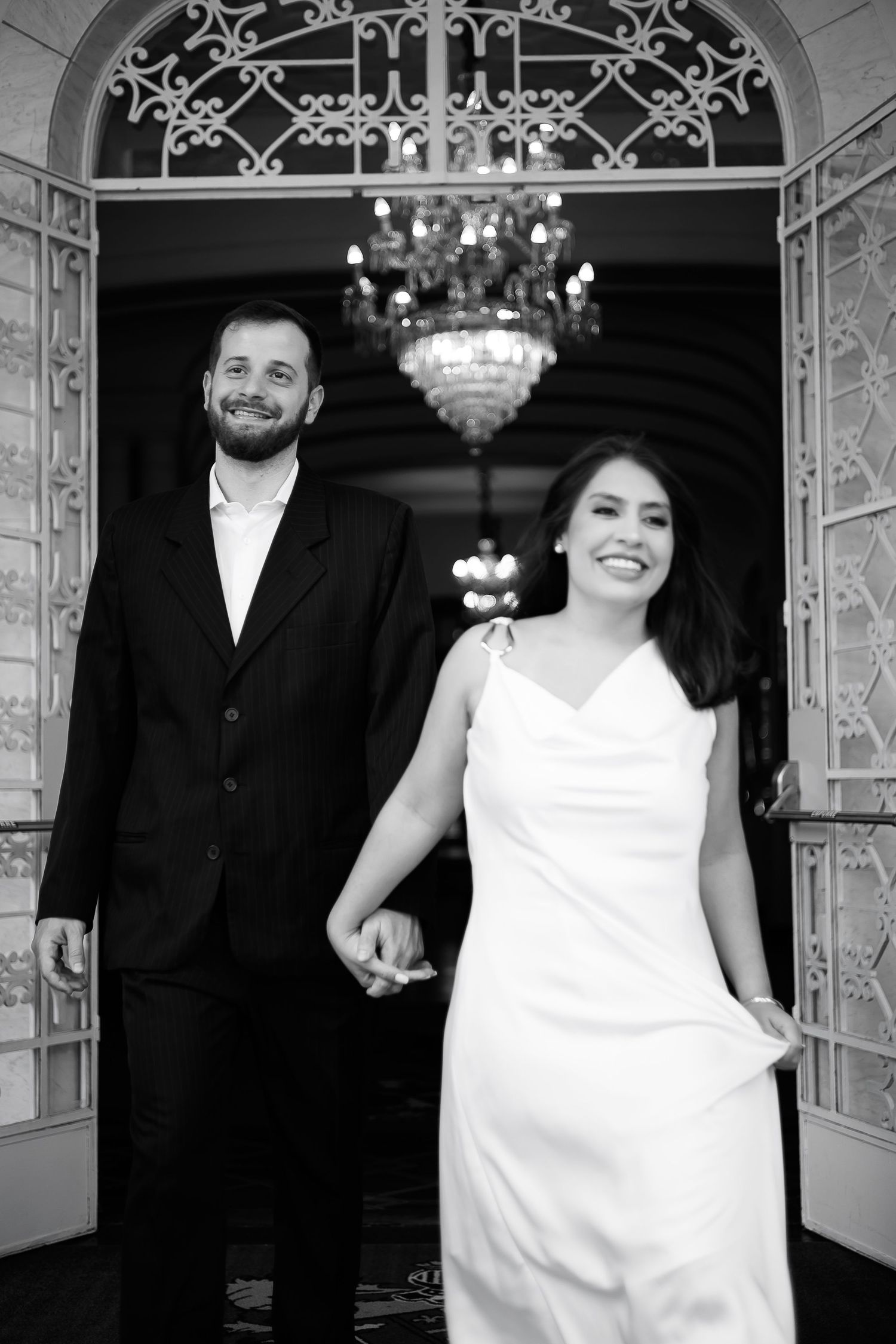 Black and white photo of smiling bride in white gown and groom in black suit holding hands outside elegant venue.