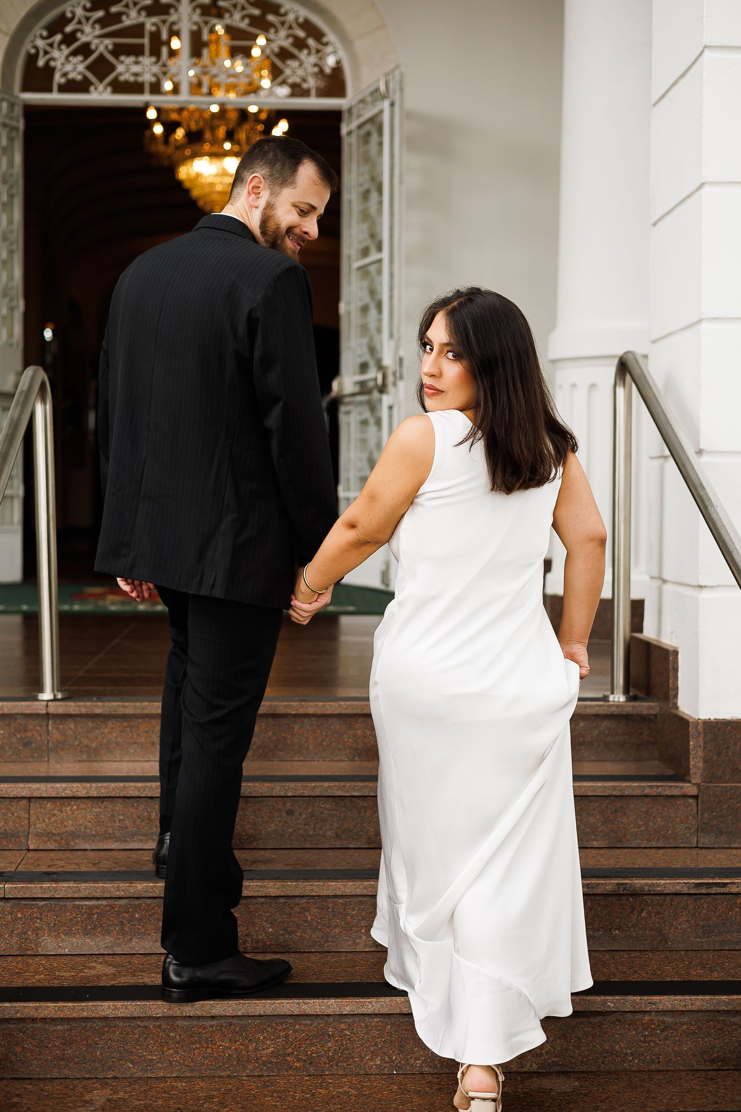 Couple in formal attire holding hands on elegant staircase, woman in white dress looks back, man in black suit smiles.