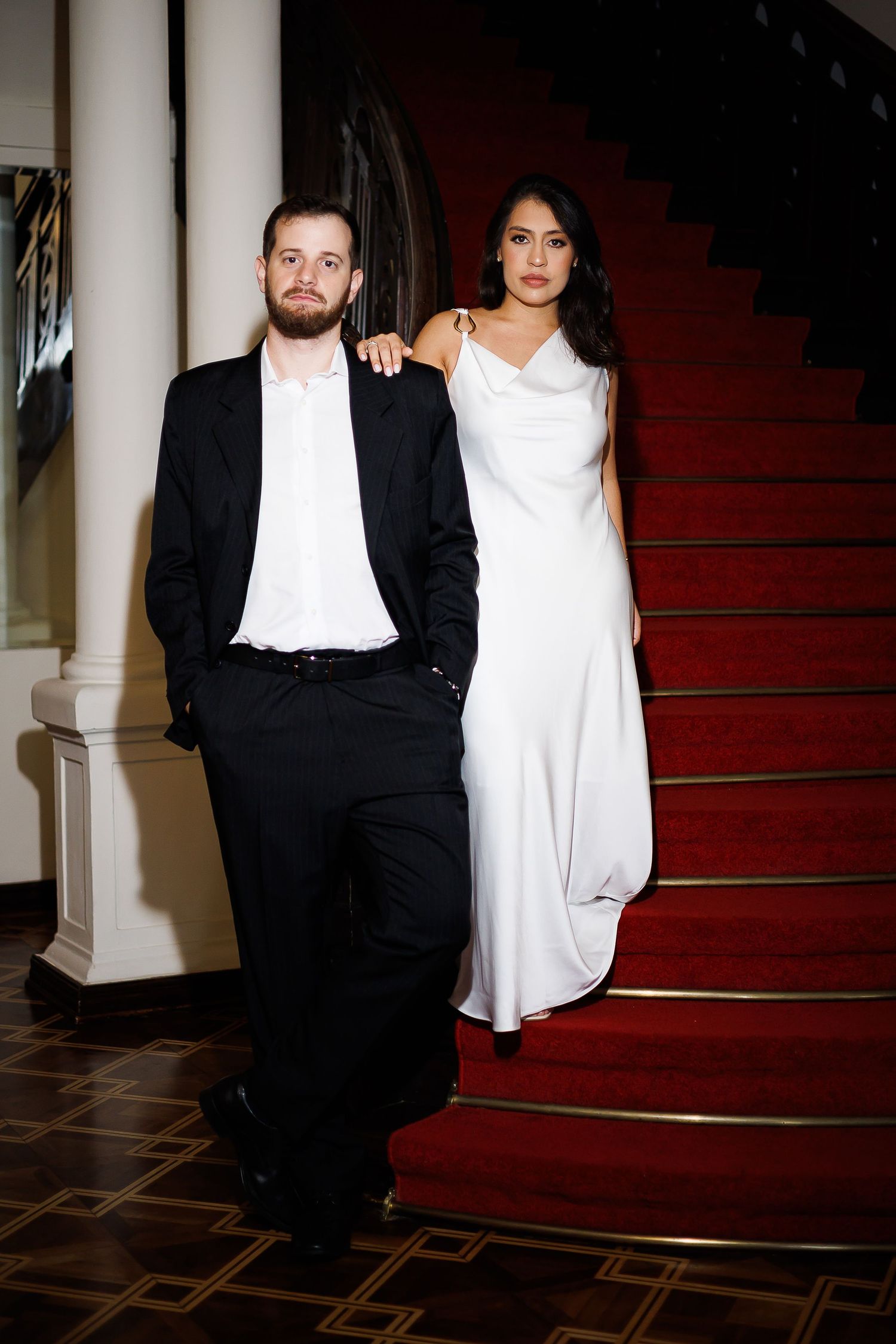 Elegant couple posing on red carpeted staircase, man in black suit, woman in white gown, formal event attire.