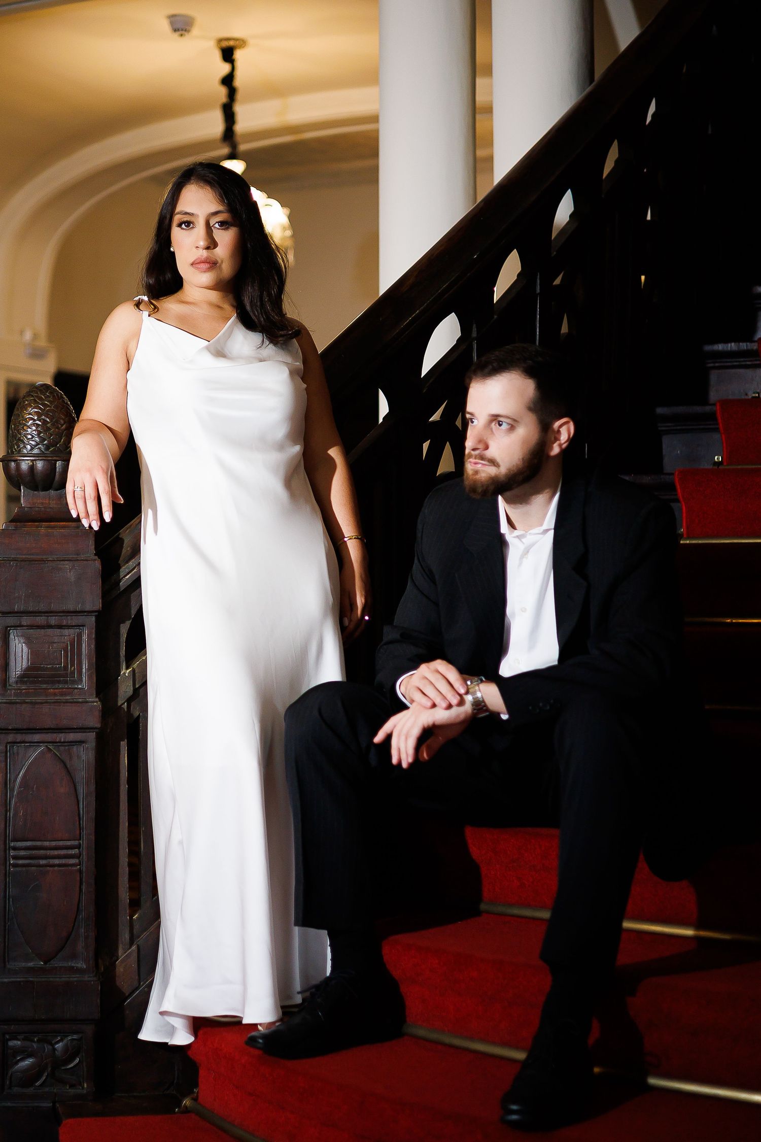 Elegant couple on red-carpeted stairs, woman in white satin gown, man in black suit, classic interior.