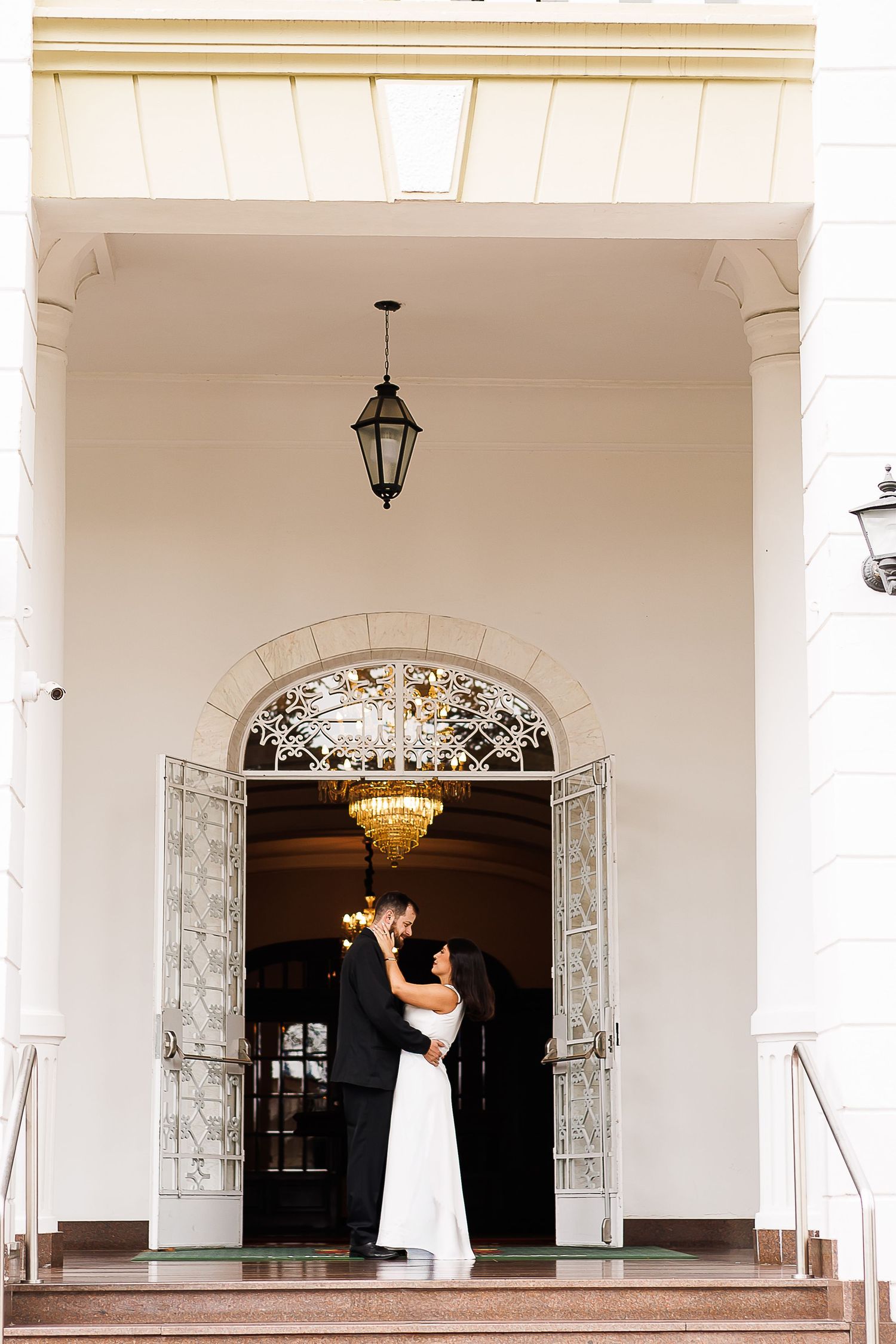 Bride and groom share romantic moment in ornate doorway with chandelier and decorative iron doors at elegant wedding venue.