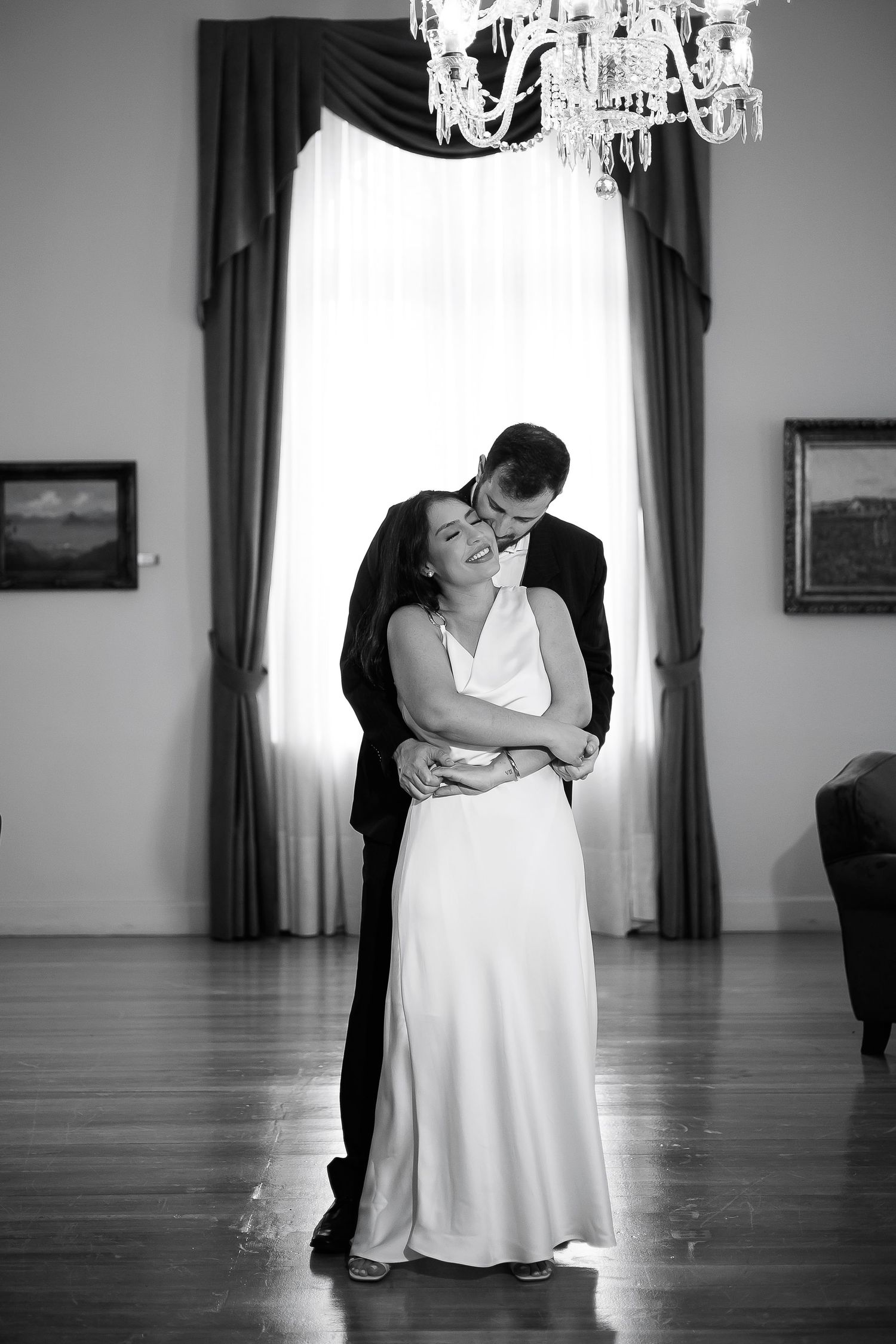 Black and white photo of a bride and groom sharing their first dance under a crystal chandelier in an elegant ballroom.