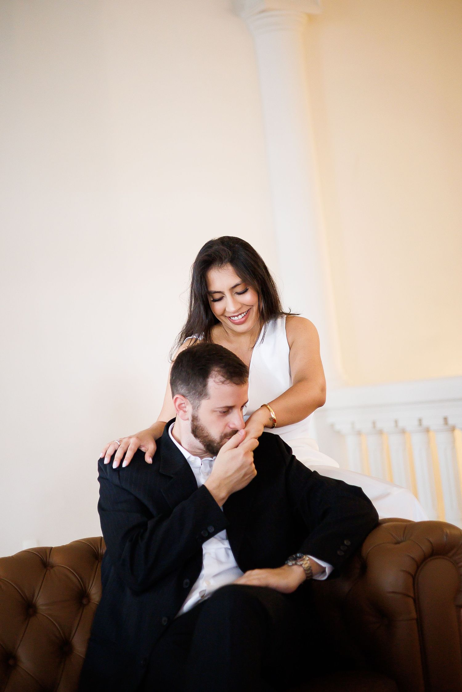 A smiling woman in a white dress stands behind a man in a black suit on a leather sofa, as he kisses her hand.