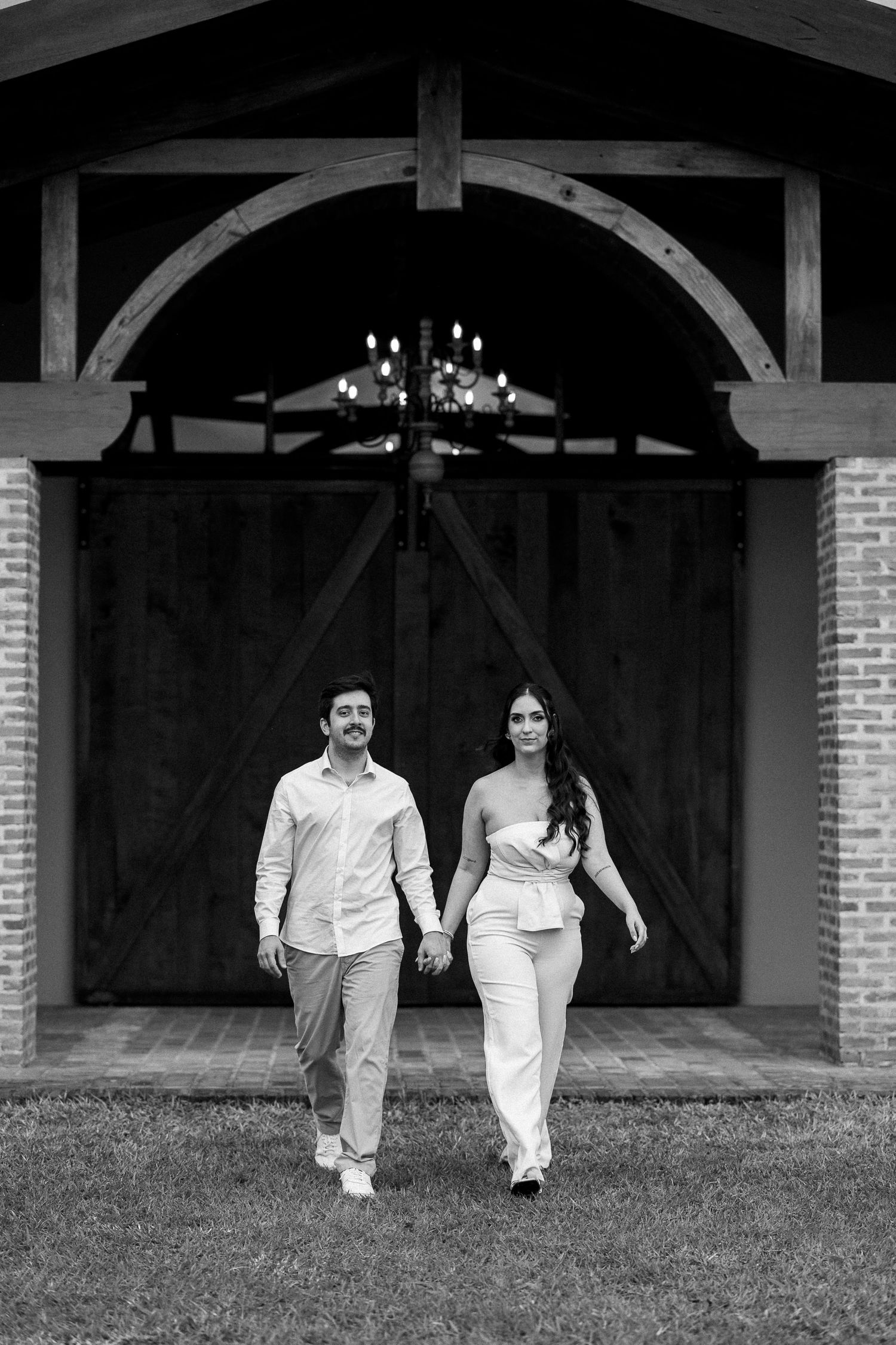 Black and white photo of a couple holding hands walking toward camera in front of rustic barn doors with chandelier.