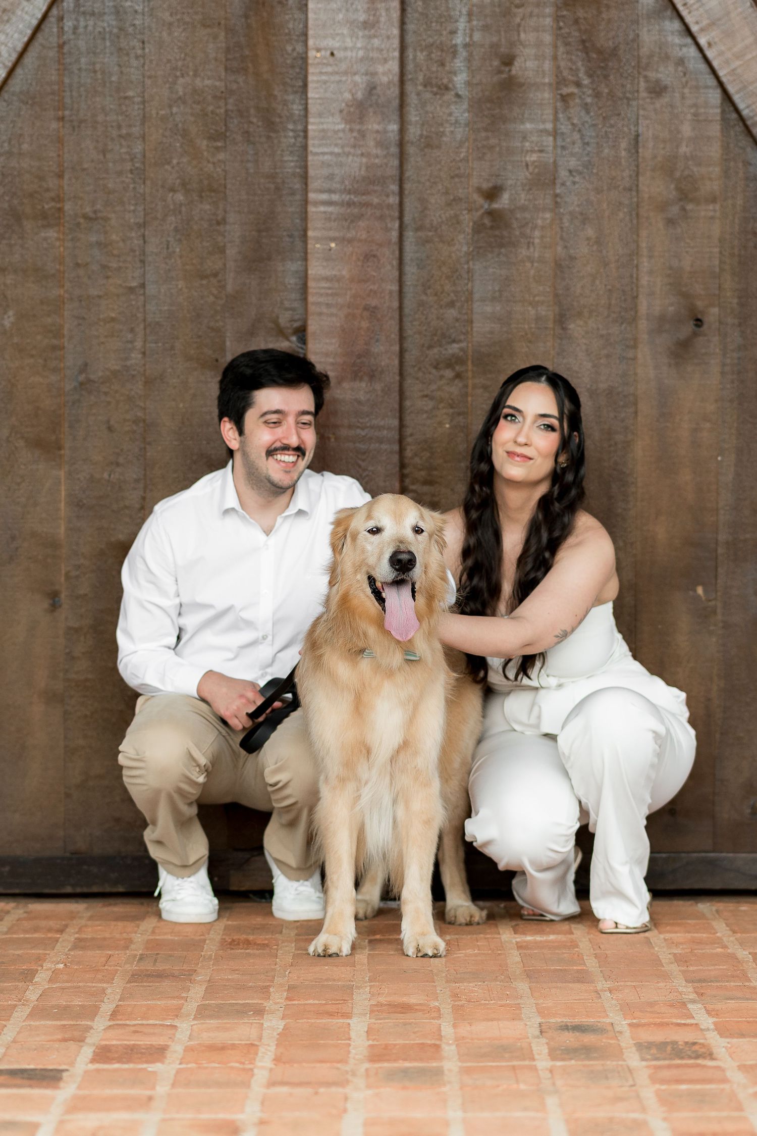 Smiling couple in white outfits crouching beside a golden retriever dog in front of rustic wooden barn doors.