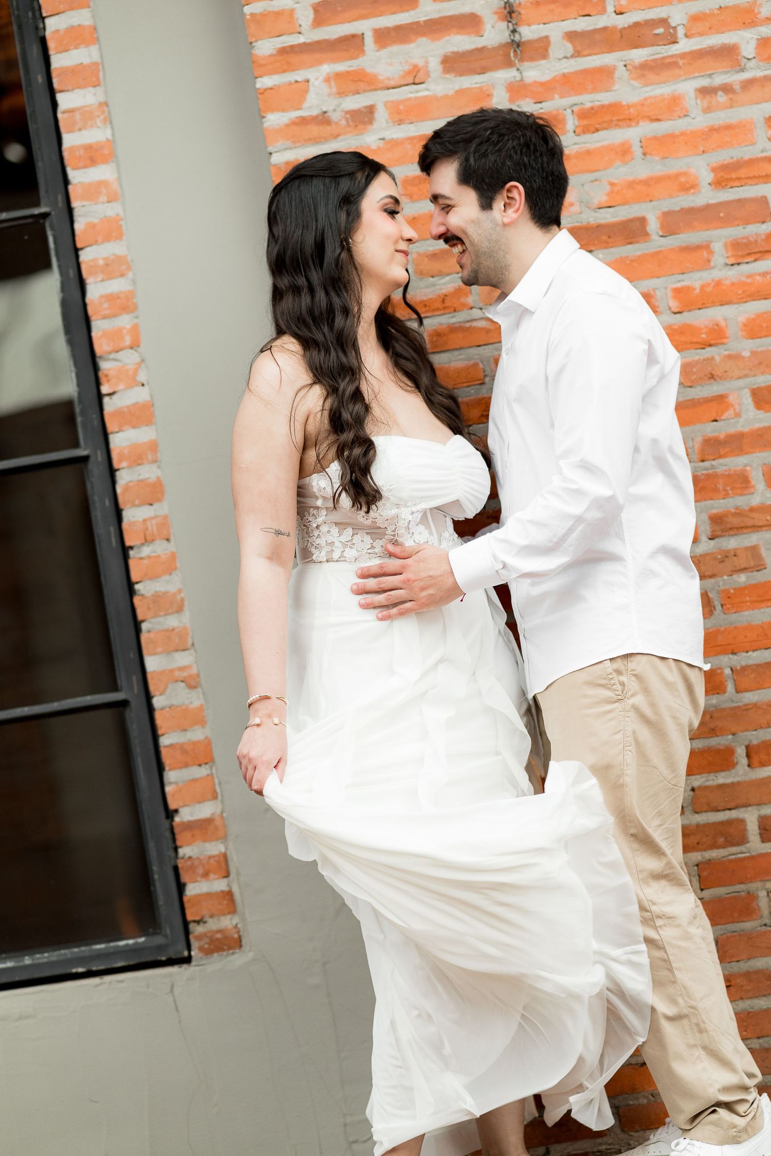Couple in white wedding attire sharing a romantic moment against a rustic brick wall outdoors.