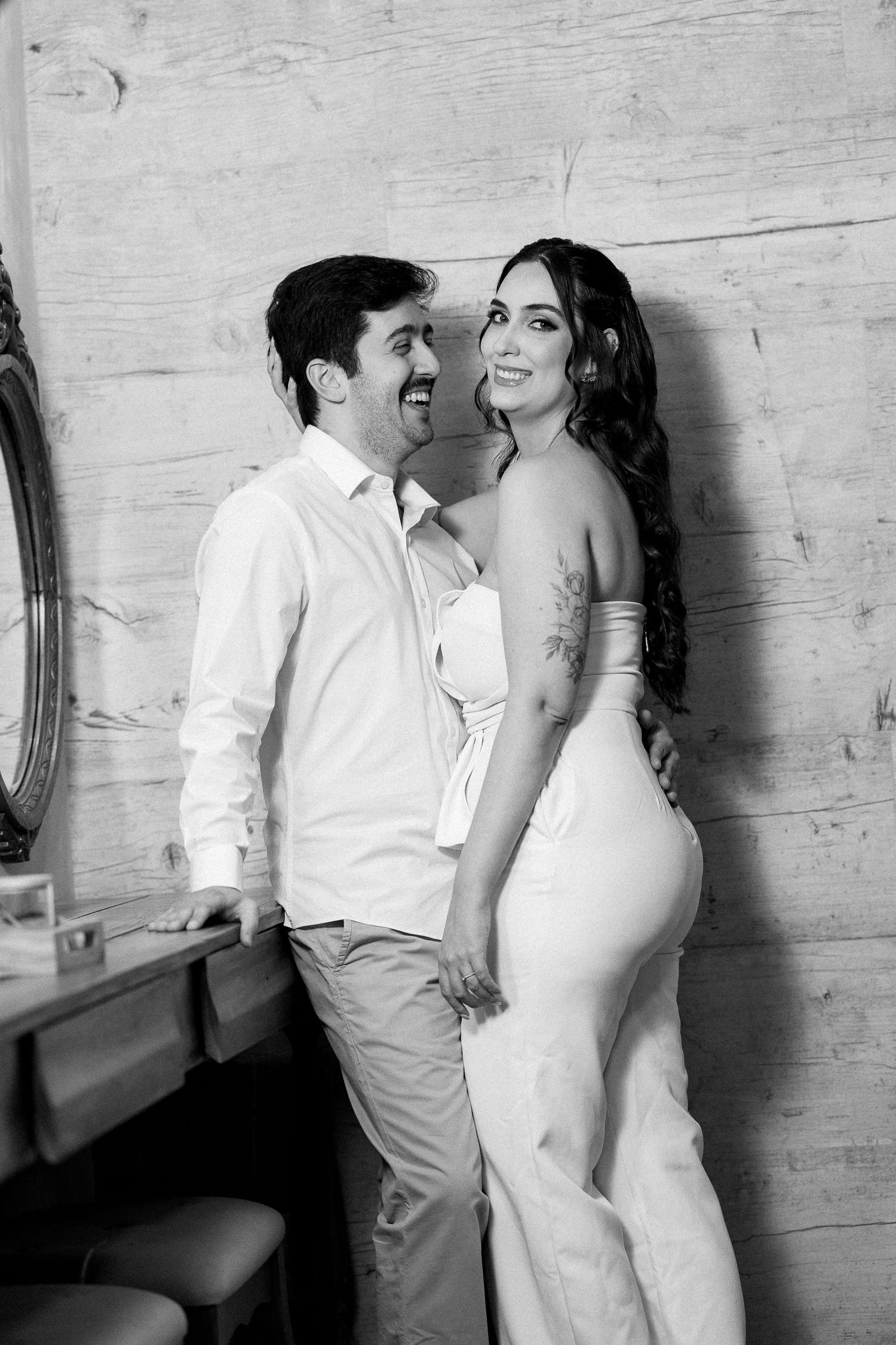 Black and white photo of a smiling couple in elegant attire posing together against a wooden wall.