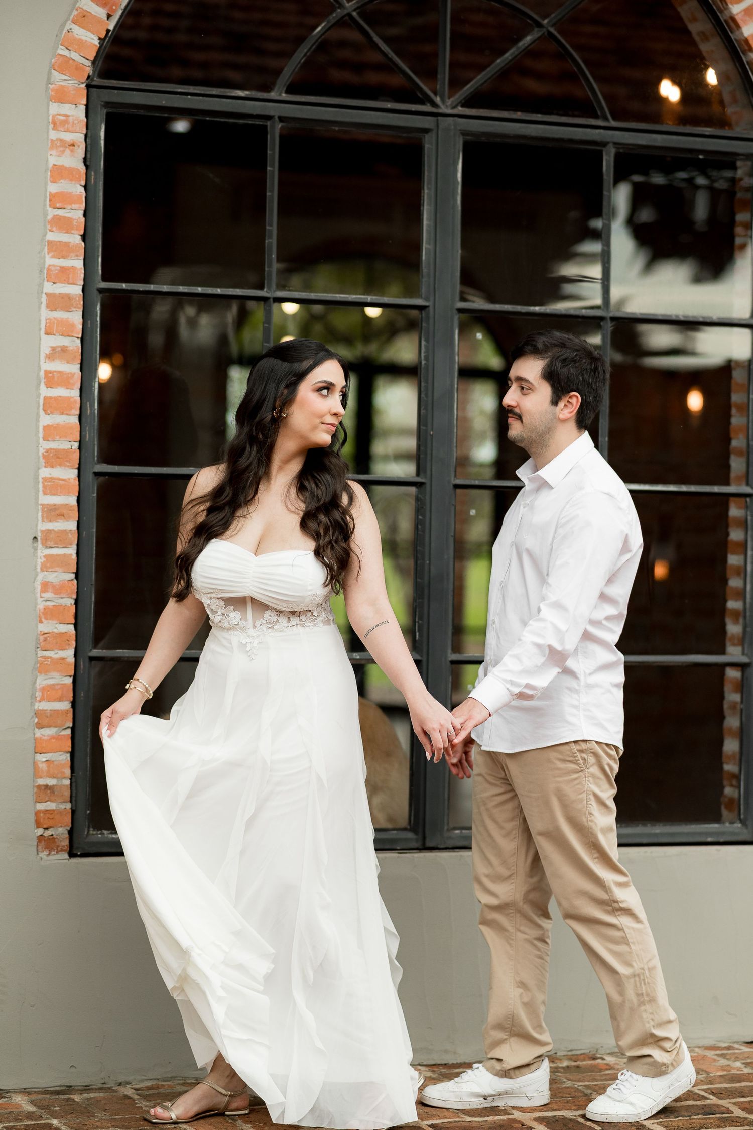 Couple holding hands in front of arched brick window, woman in white dress, man in white shirt and khaki pants.
