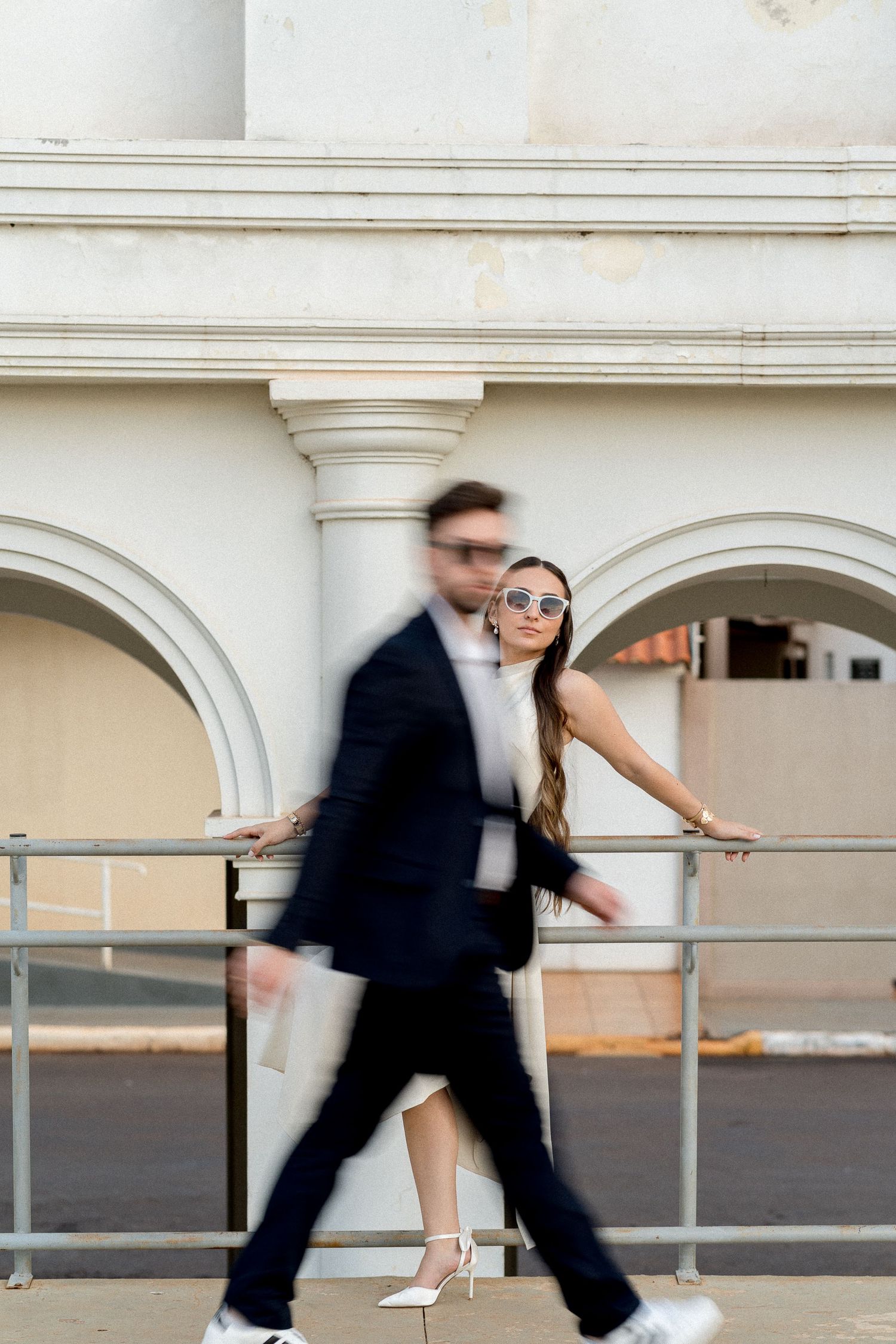 Couple posing by white arched building, man in motion blur wearing suit, woman in white dress and sunglasses.