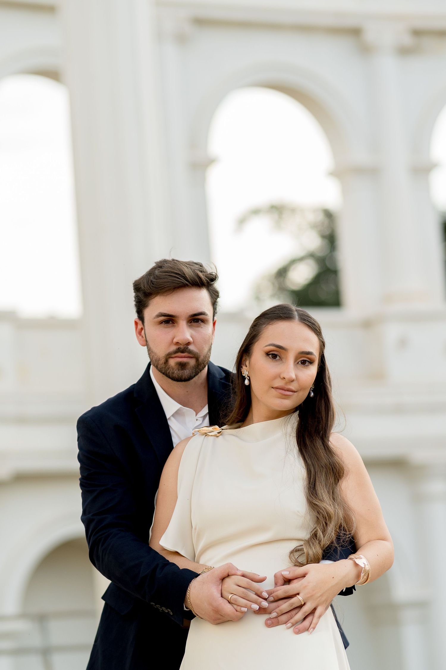 Elegant couple posing in front of white arches, man in dark suit embracing woman in cream dress.