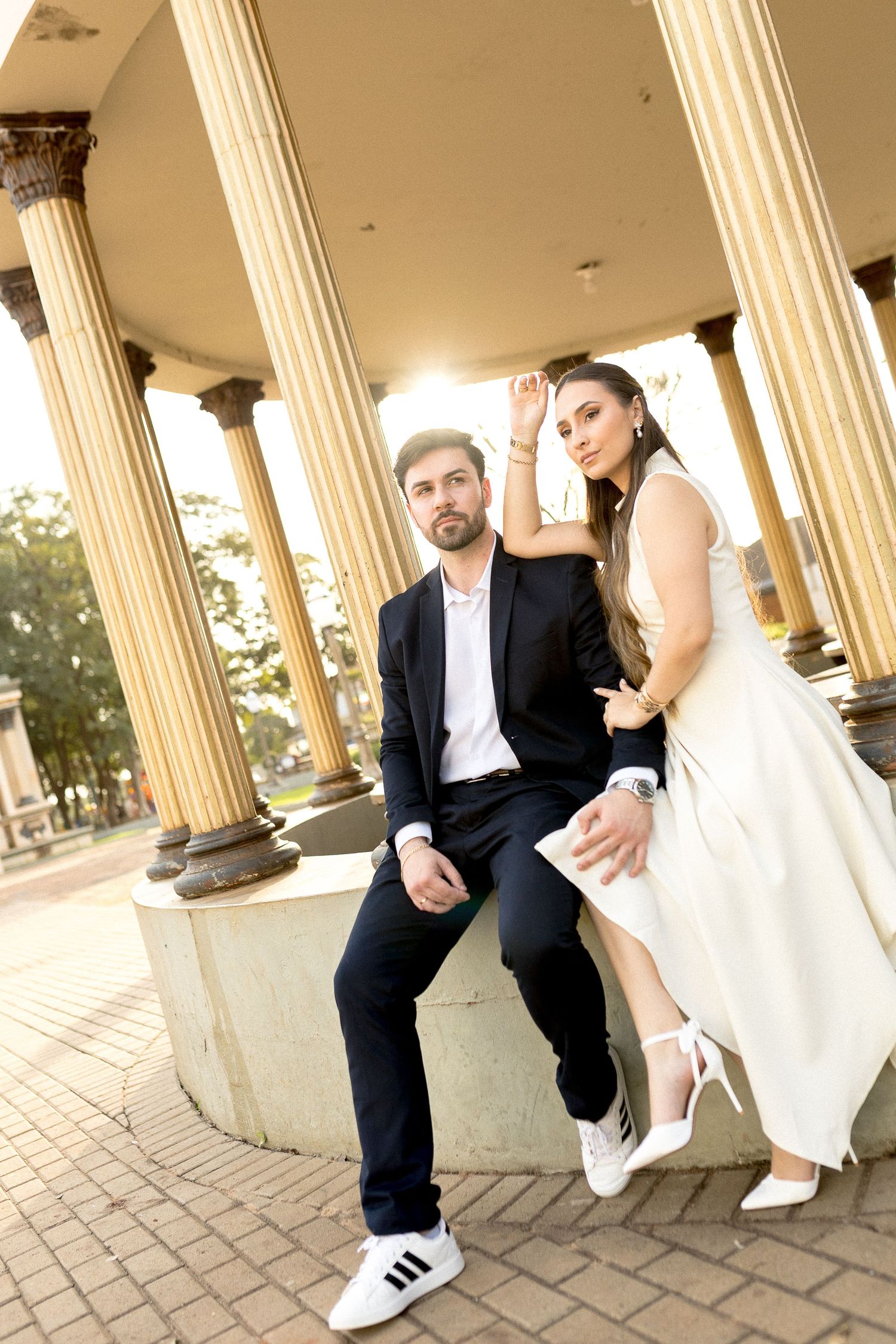 Elegant couple posing at a classical colonnade, man in black suit seated, woman in white dress standing beside him.