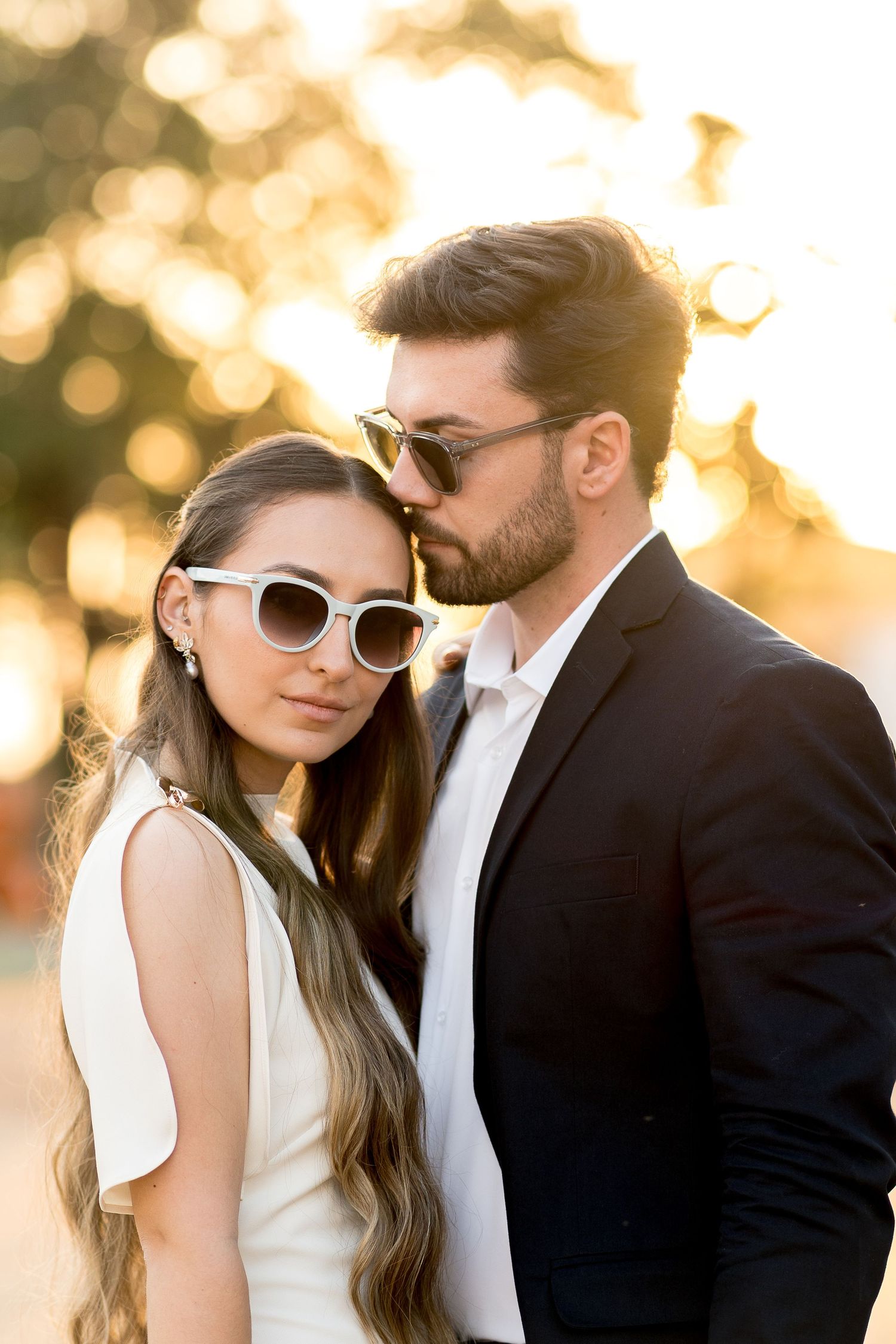 Stylish couple in sunglasses posing outdoors at golden hour, woman in white dress, man in black suit.
