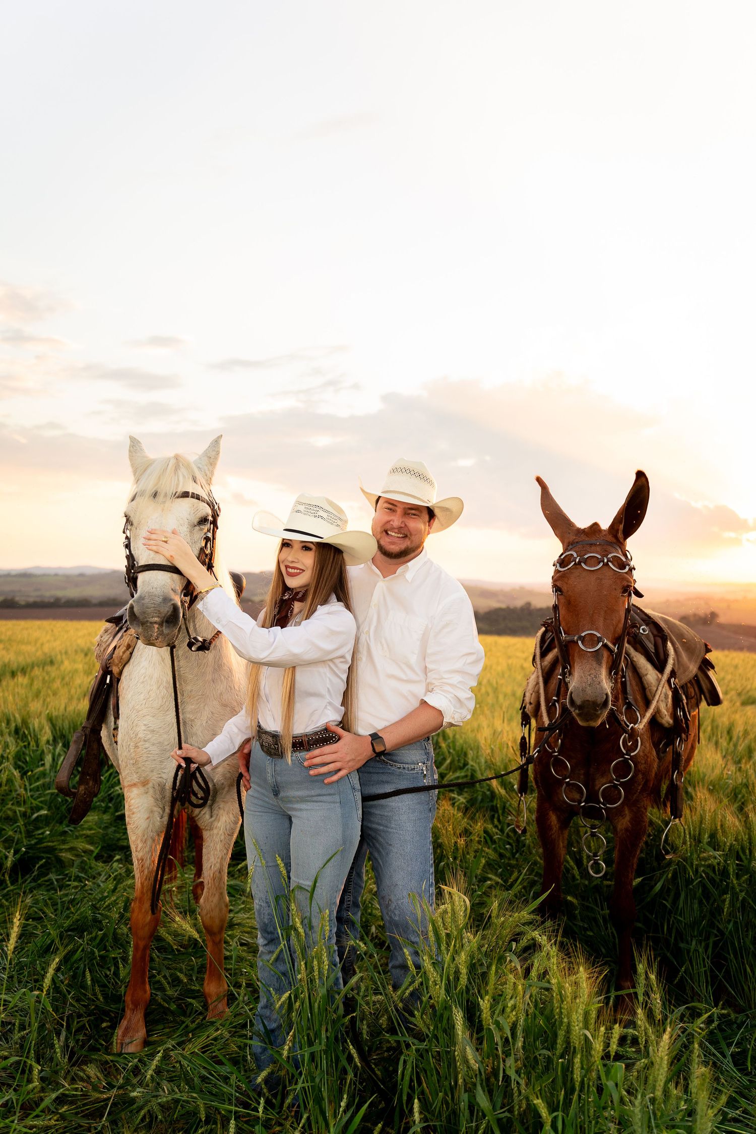 A couple in cowboy hats and jeans pose with two horses in a green field at sunset.