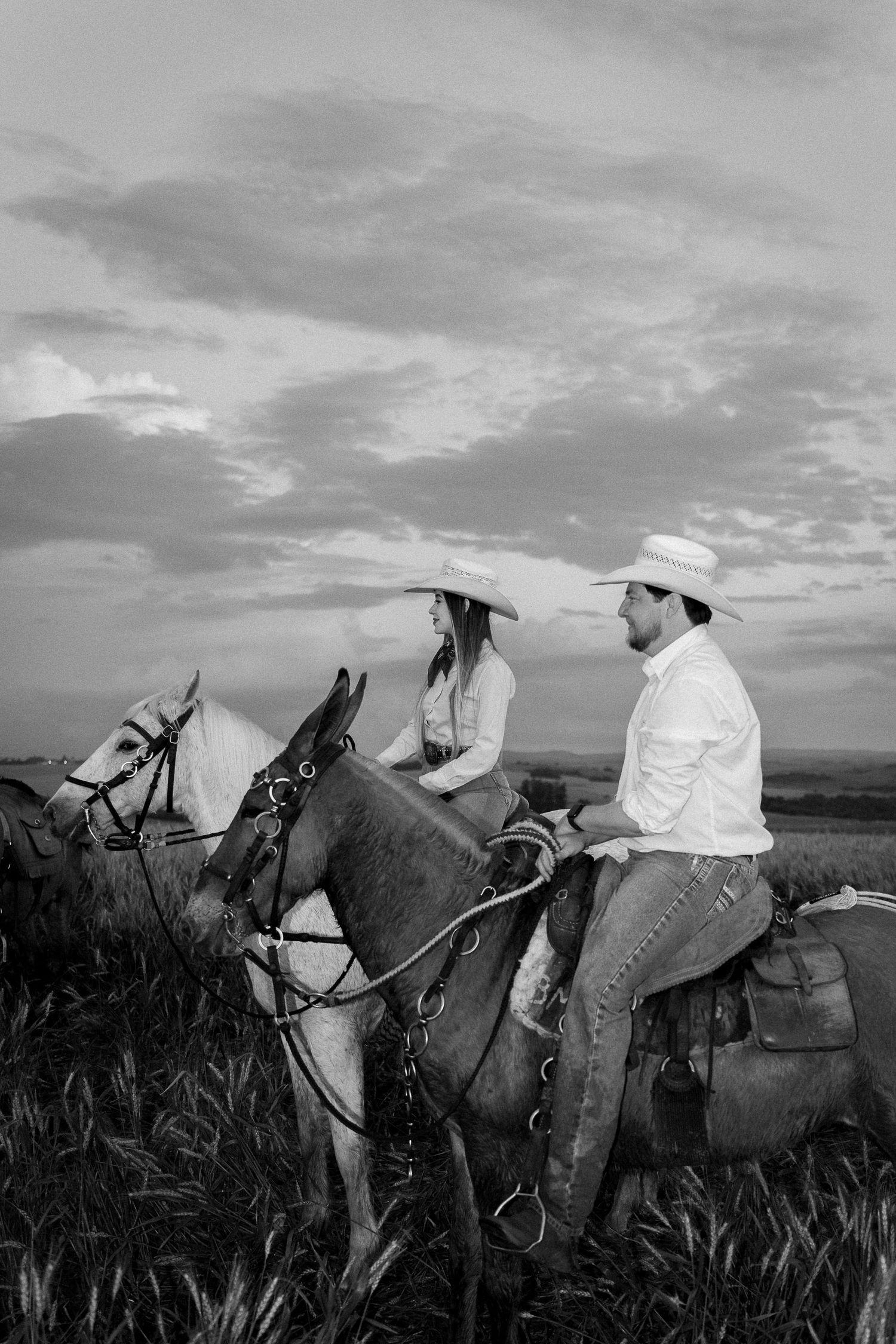 Black and white photo of a couple in cowboy hats riding horses in a field at sunset.