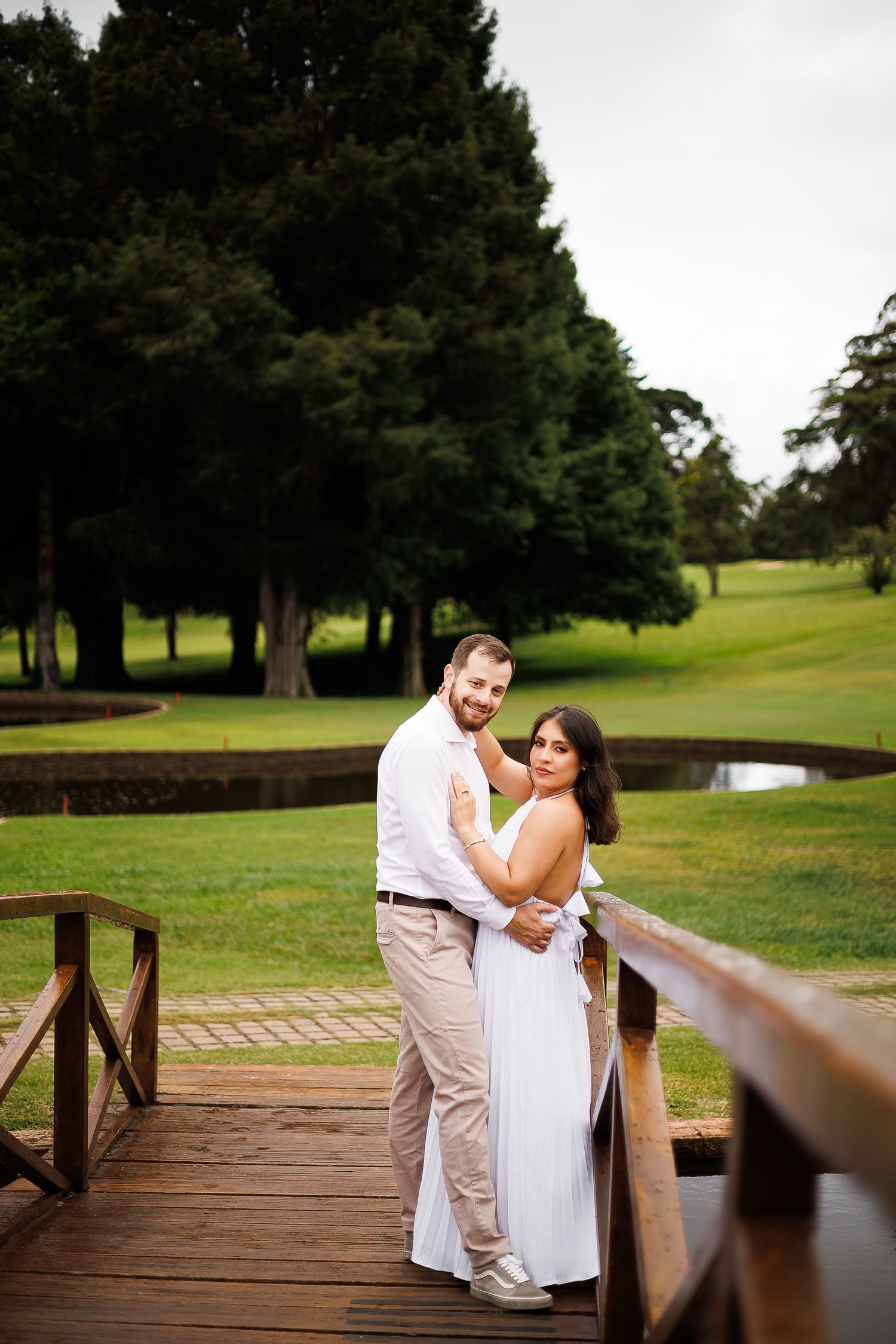 Couple in white attire embracing on a wooden bridge surrounded by lush green trees and manicured lawn.