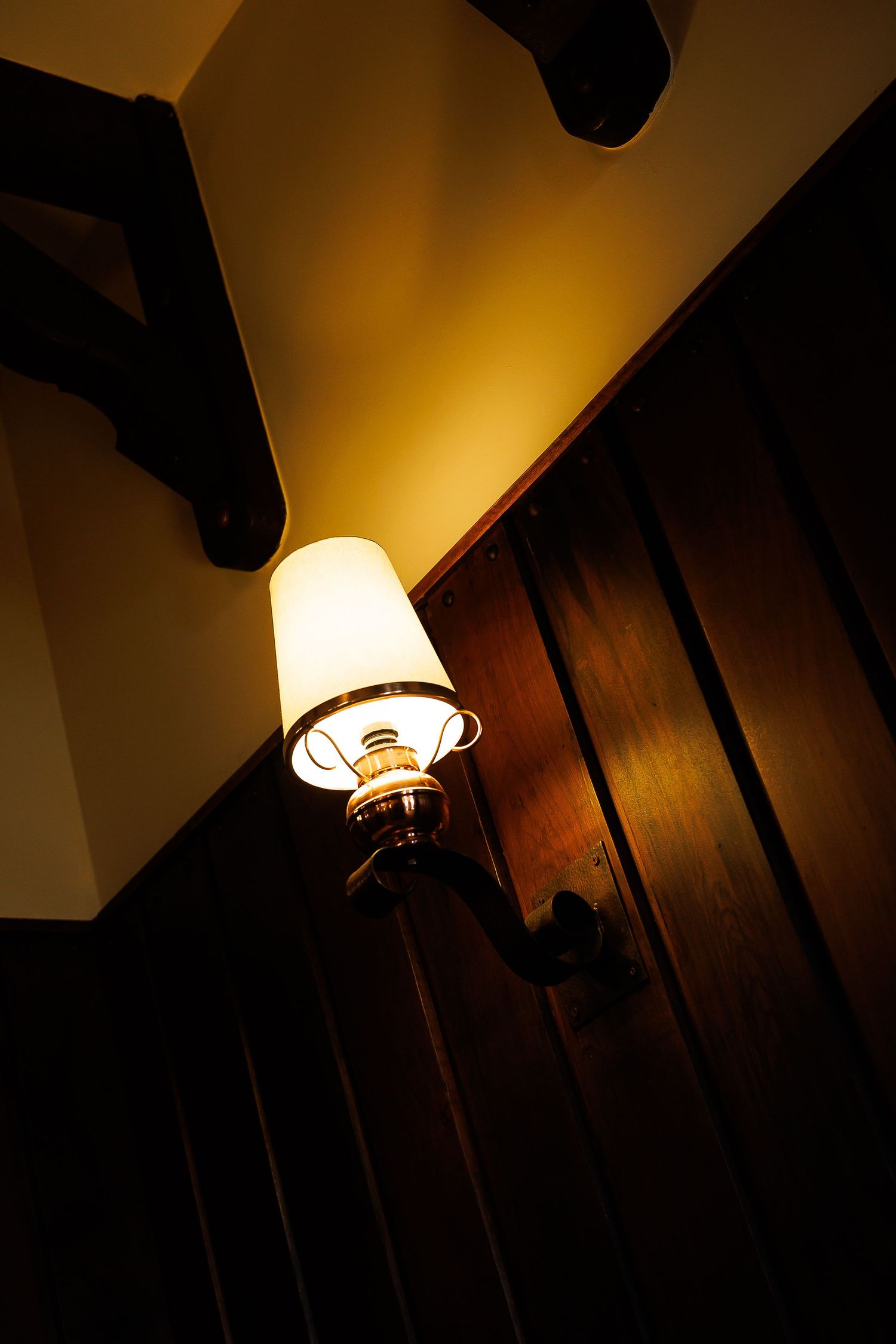 Vintage wall-mounted oil lamp glowing warmly against dark wood paneling near a staircase.