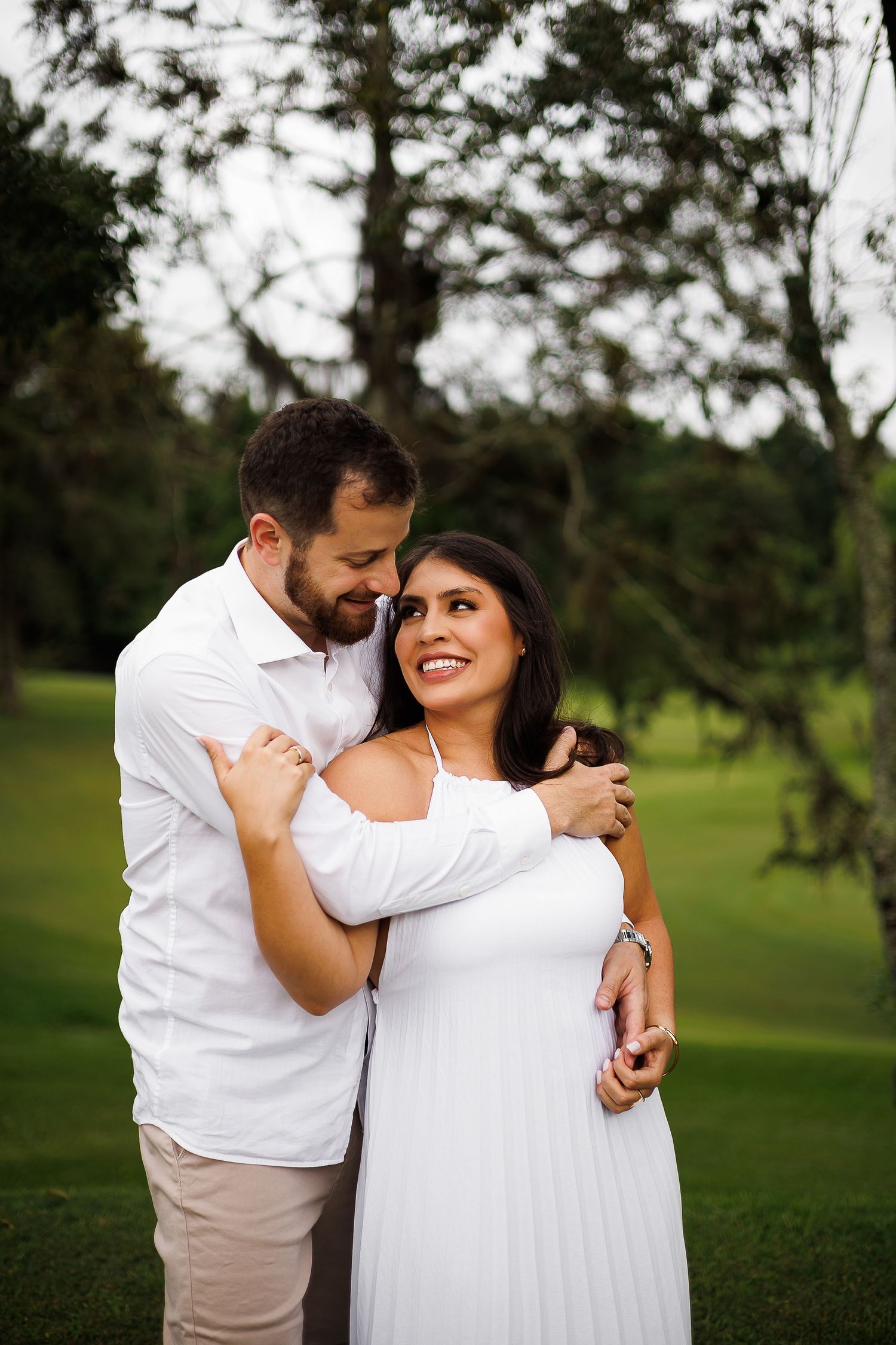 A smiling couple in white outfits embrace outdoors on a green lawn with trees in the background.