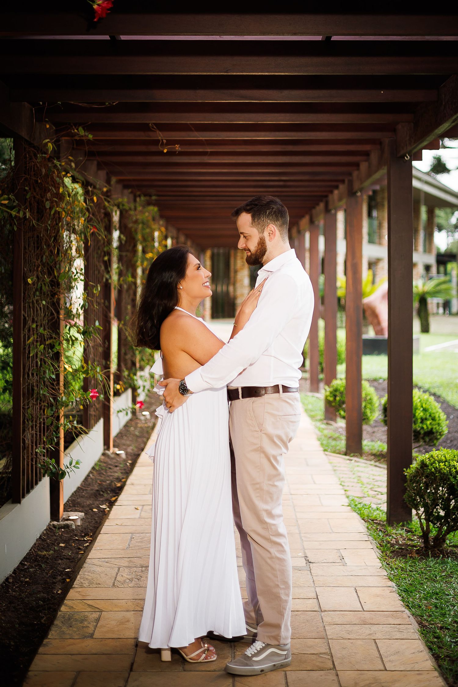 A couple in white attire embraces under a wooden pergola surrounded by lush greenery.