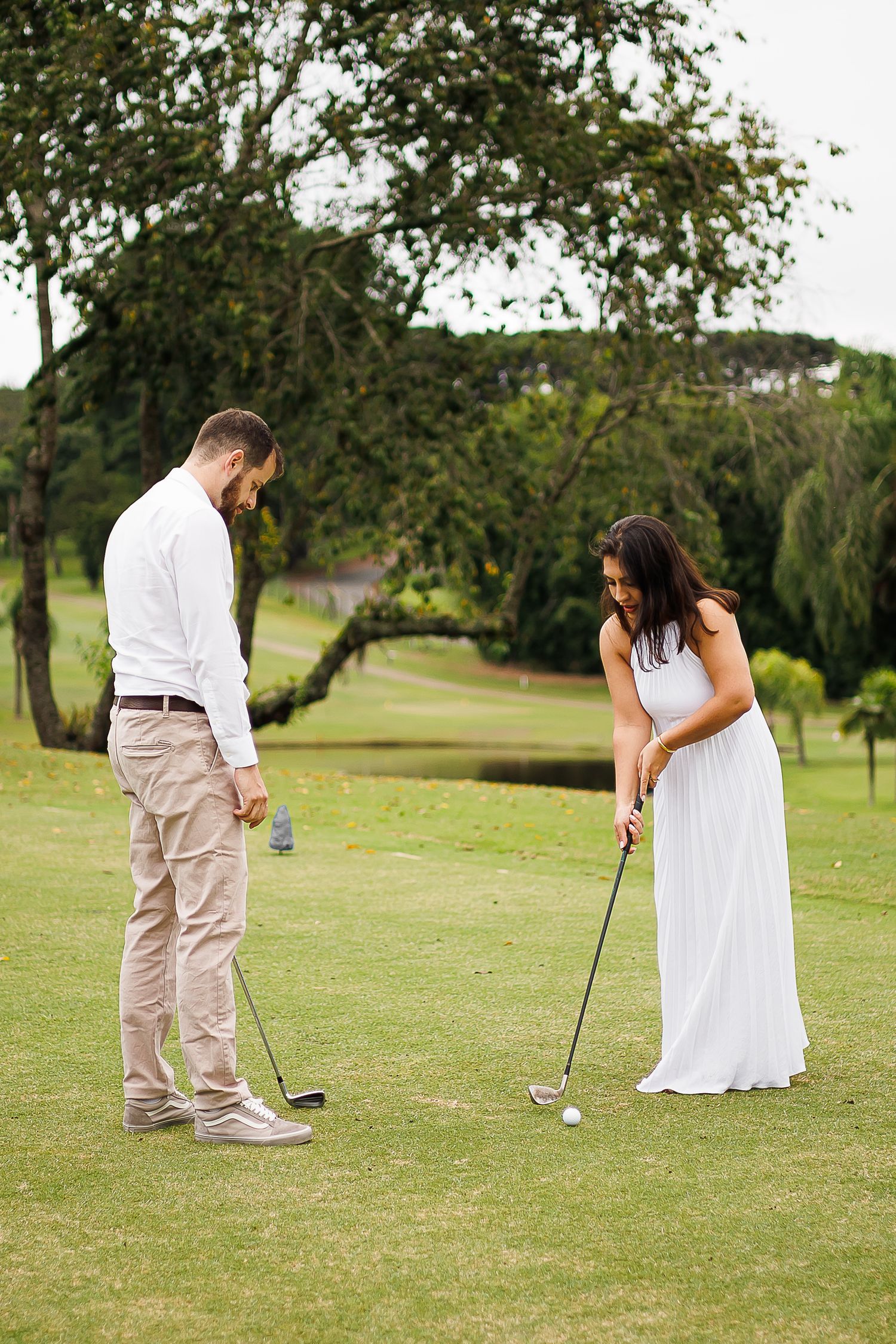 Couple in white wedding attire playing golf on a lush green course surrounded by trees.
