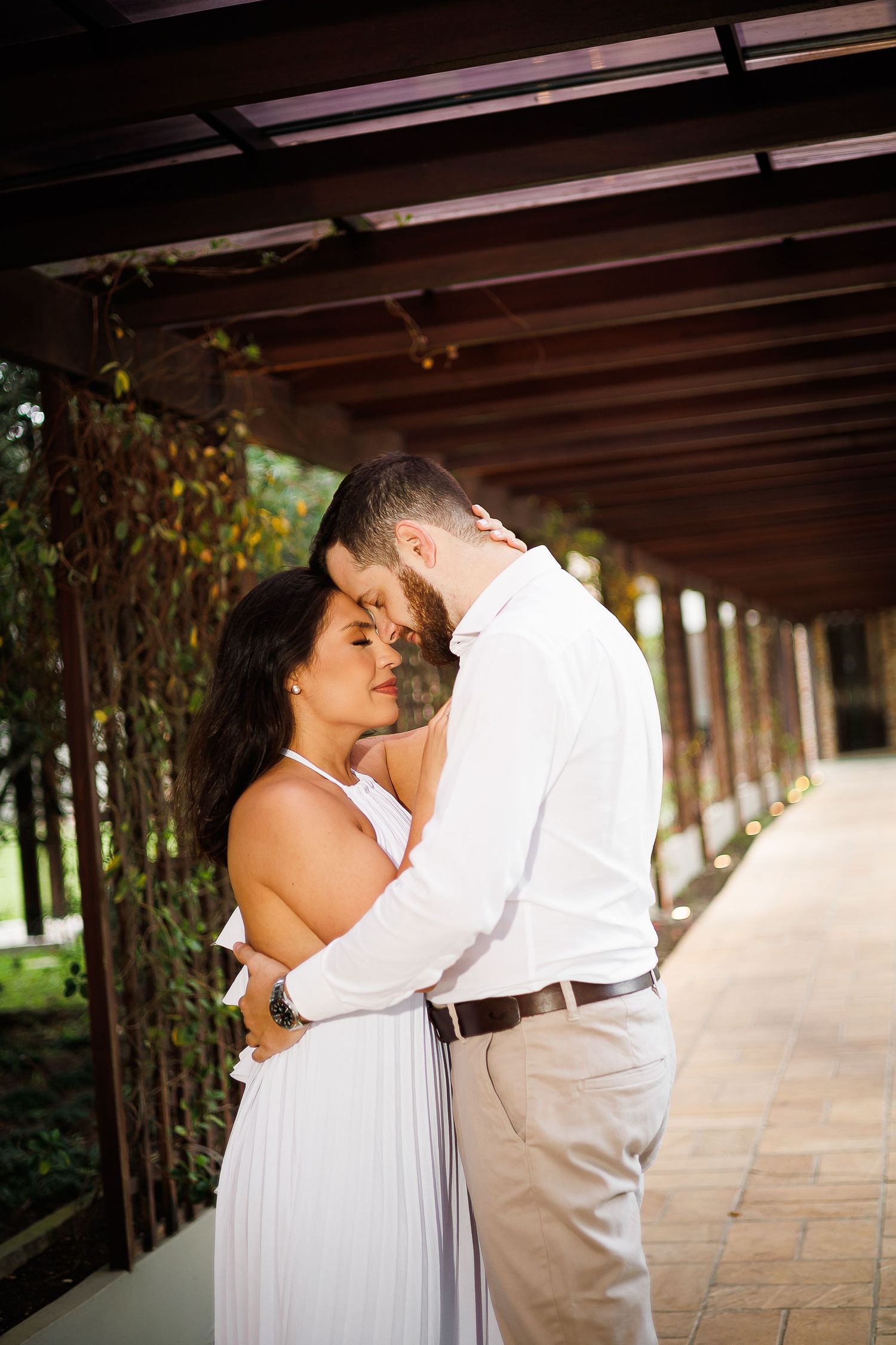 Couple embracing tenderly under a wooden pergola, woman in white dress, man in white shirt and khaki pants.