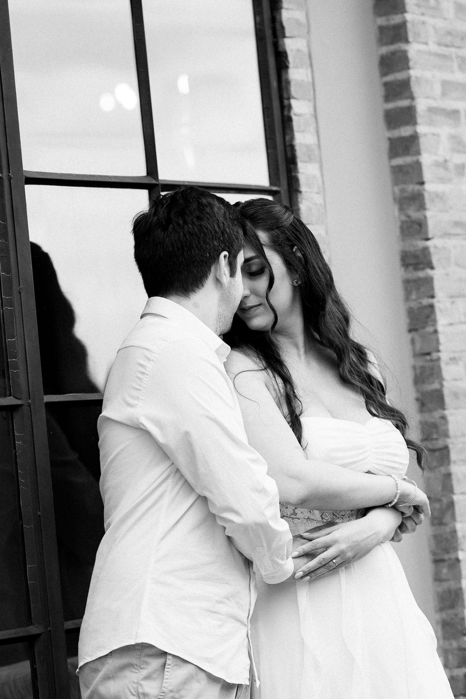 Black and white photo of a couple embracing tenderly near a brick wall and large window.