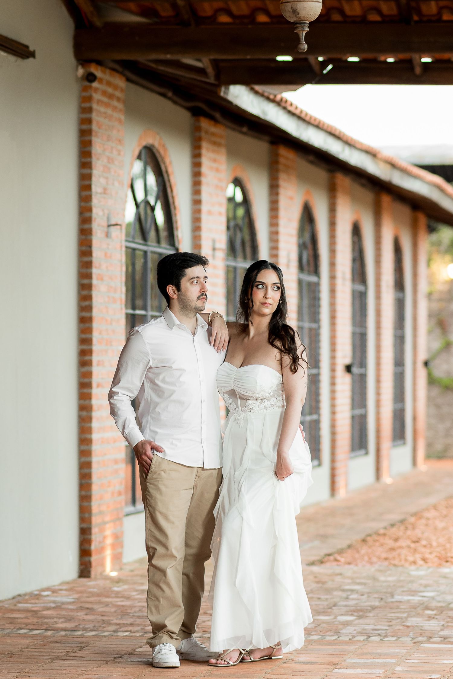 Couple in white attire posing together on a brick walkway outside an elegant building with arched windows.