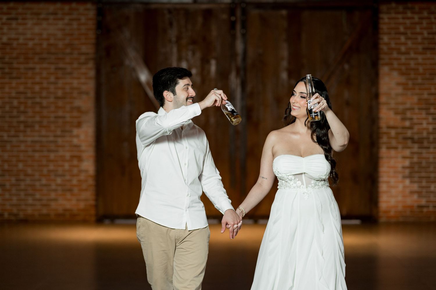 Bride and groom holding hands while drinking champagne bottles in a rustic brick barn wedding venue.