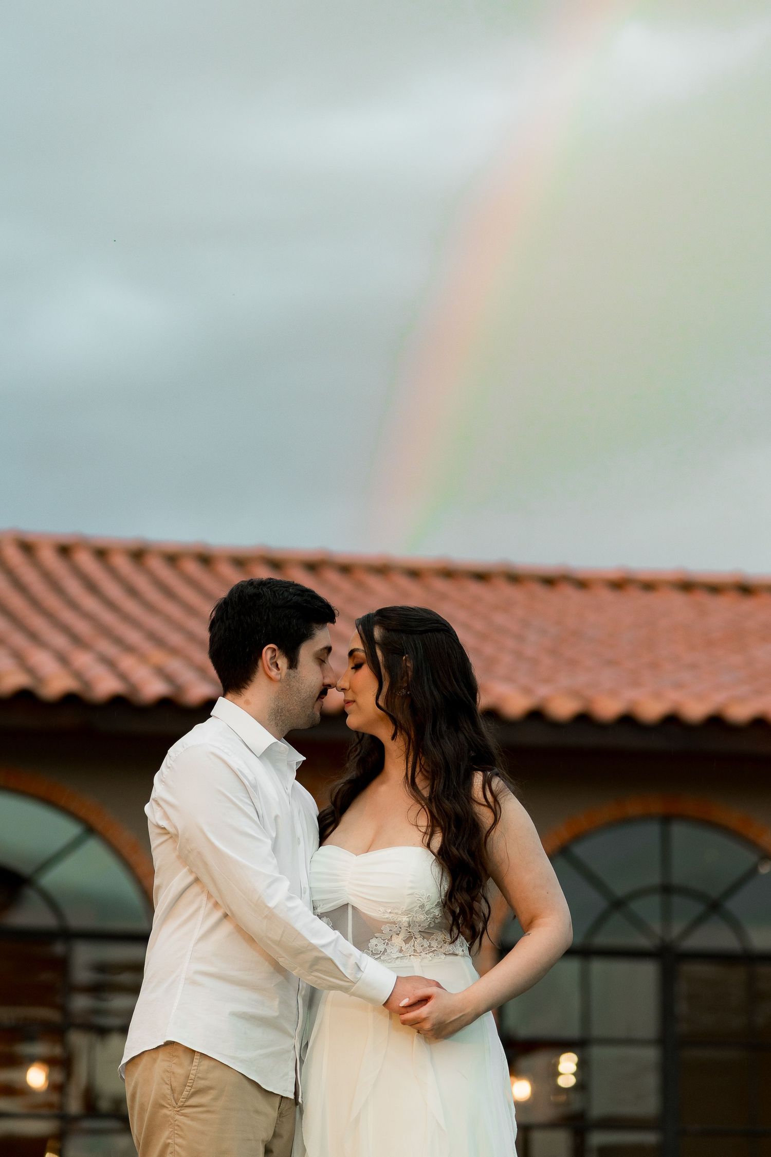A couple in wedding attire shares a romantic moment outdoors with a rainbow arching over a terracotta roof behind them.