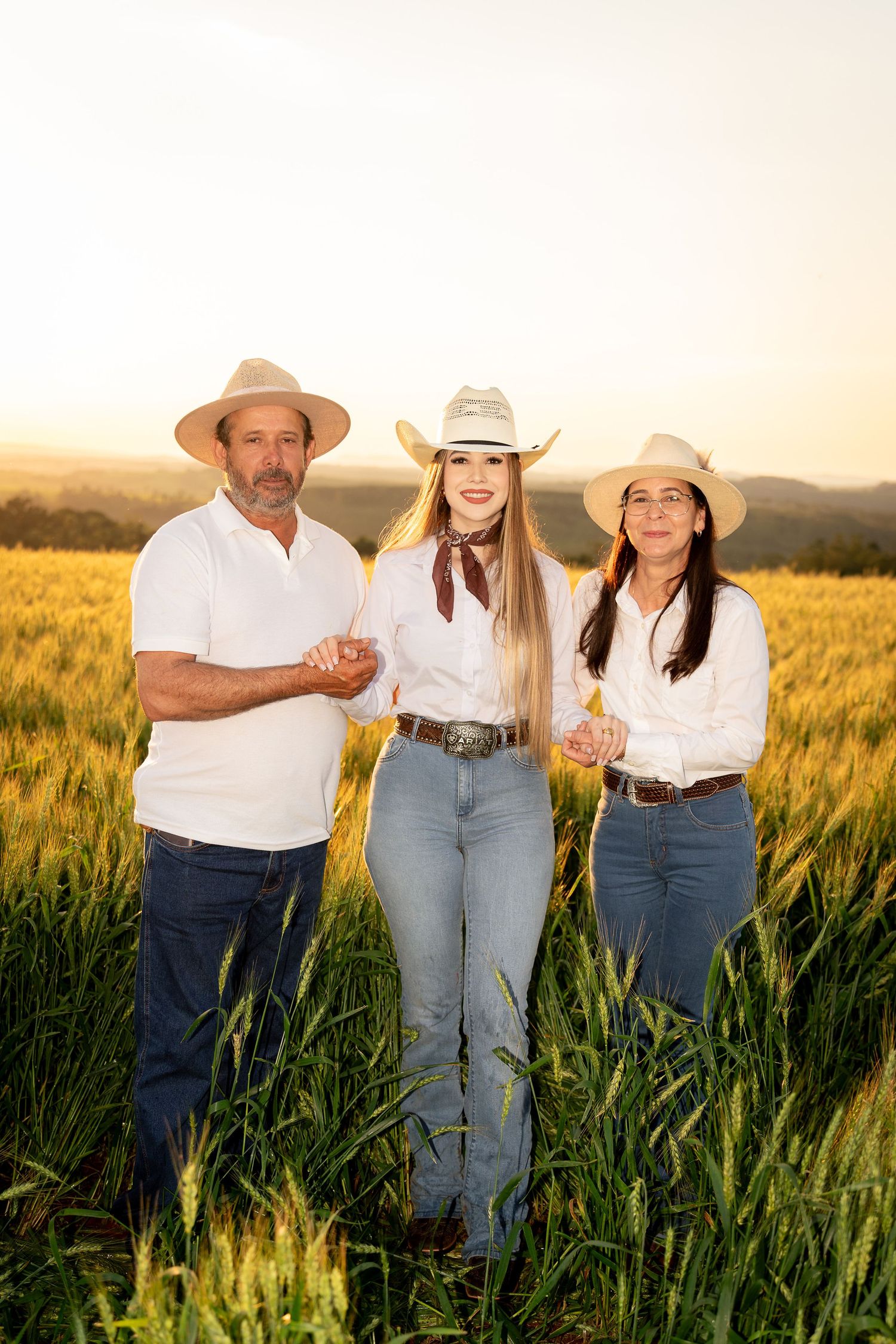 Three farmers in cowboy hats and white shirts stand smiling in a golden wheat field at sunset.