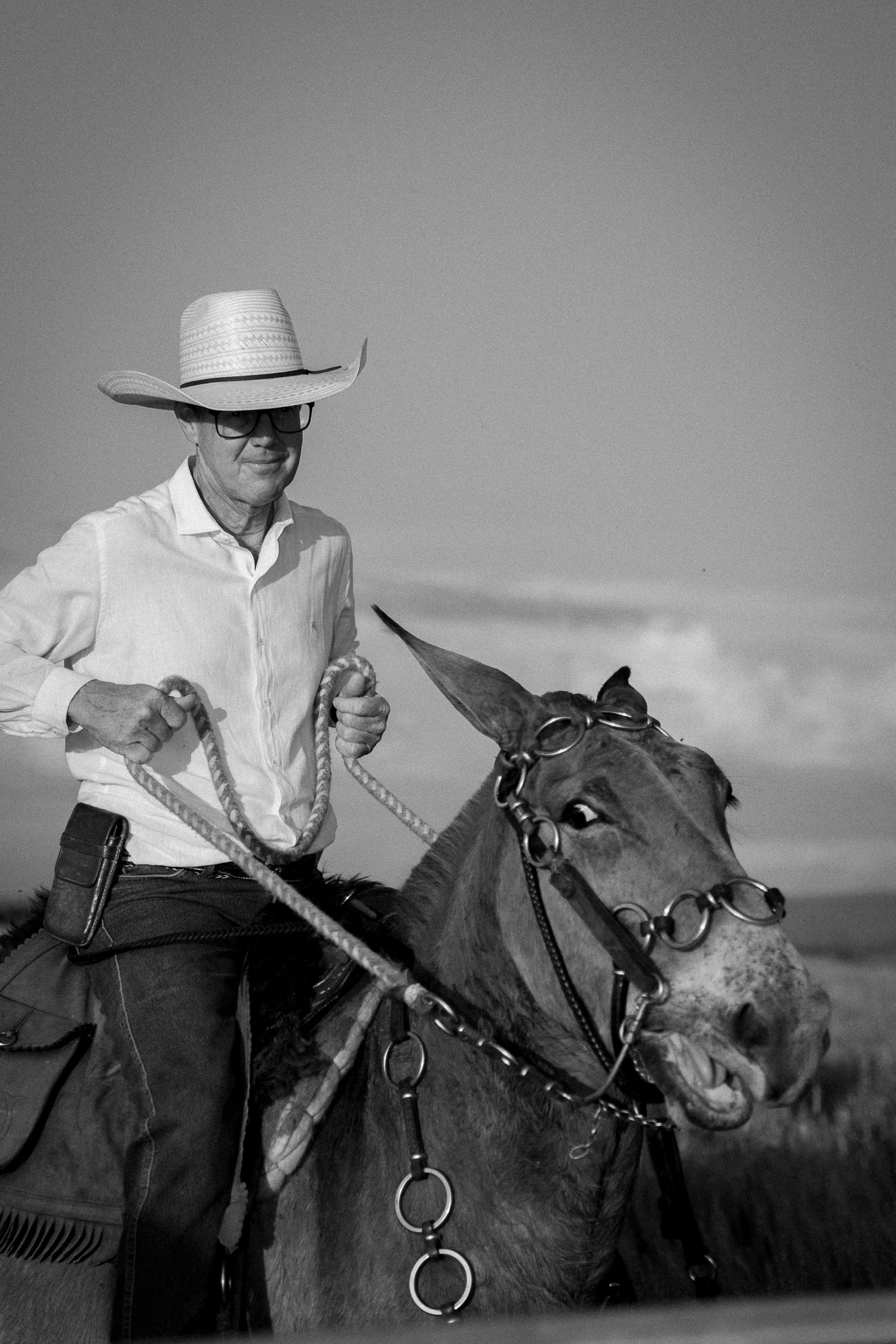 Black and white photo of a cowboy in white shirt and hat riding a mule with rope reins in an open field.