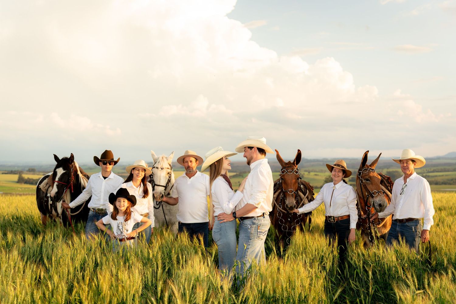 A family in white shirts and cowboy hats poses with horses in a lush green field under a cloudy sky.
