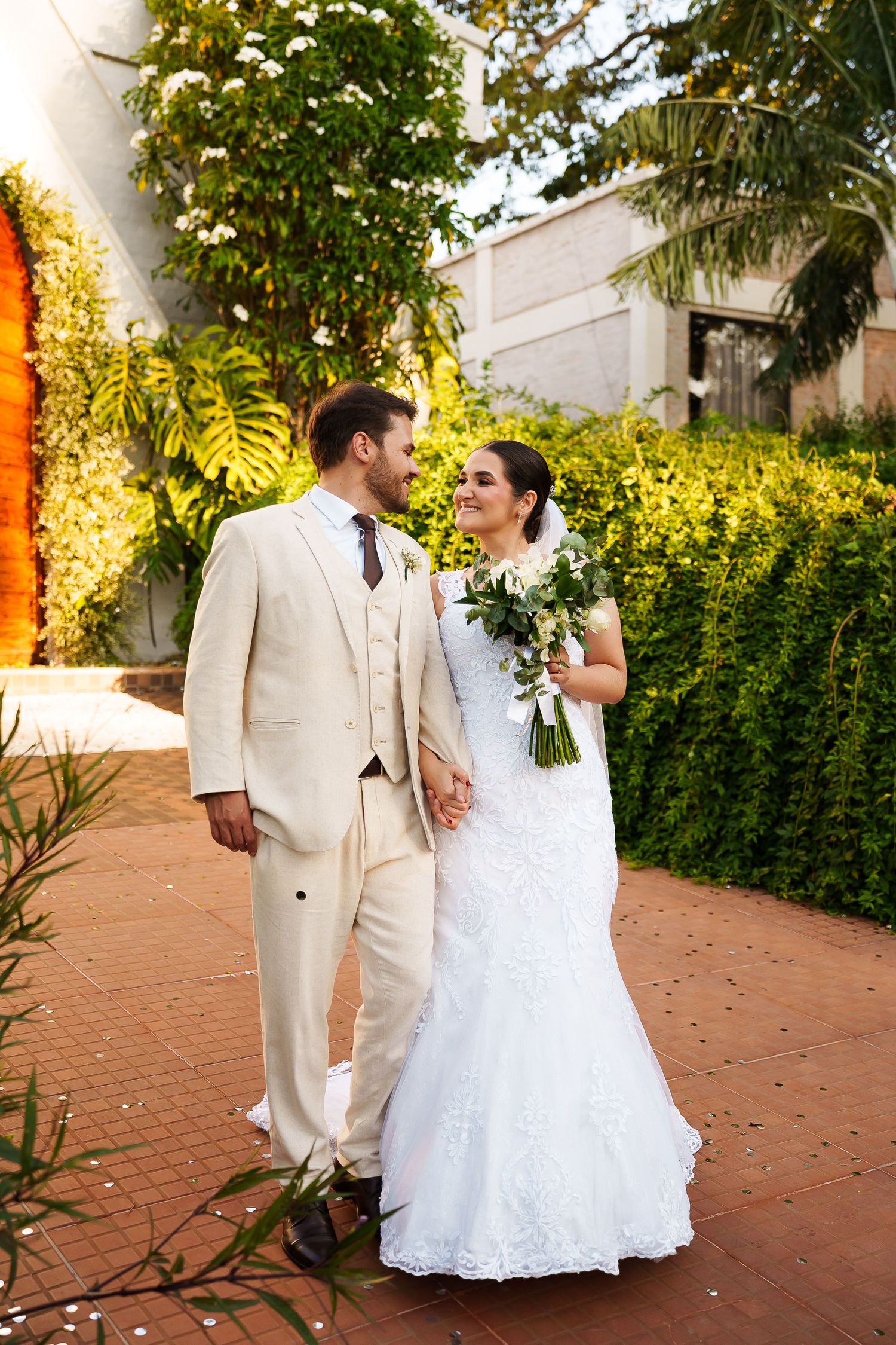 Bride and groom walk hand in hand outdoors, she holds white bouquet in lace gown, he wears cream suit.