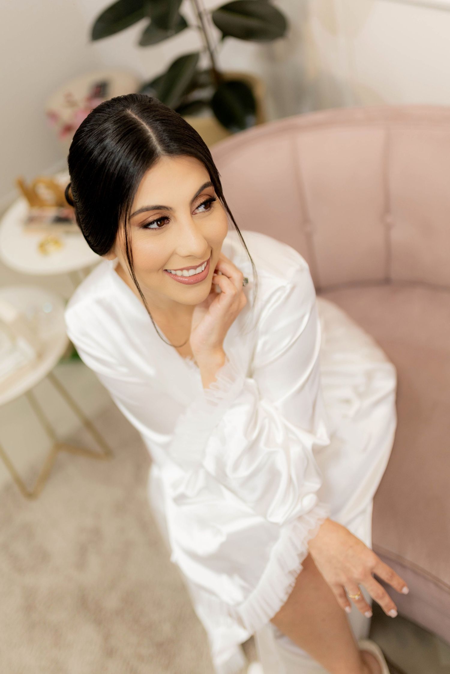 Smiling bride in white satin robe sitting in pink chair, with elegant updo and natural makeup.