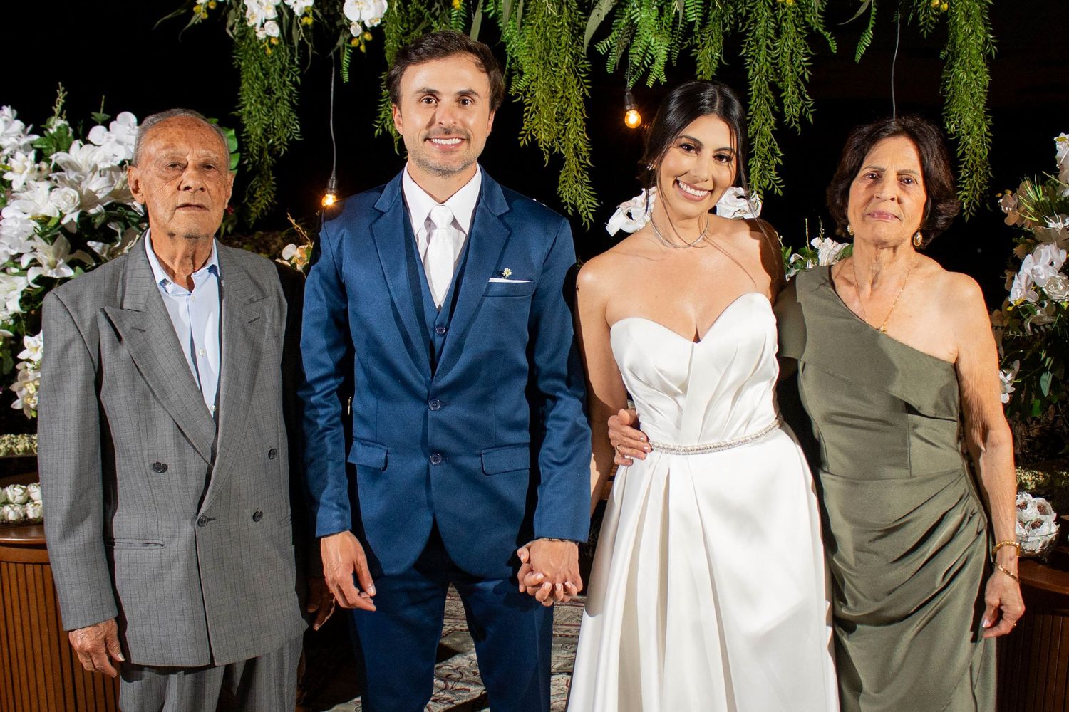 Bride and groom pose with elderly family members at elegant wedding ceremony with floral backdrop.