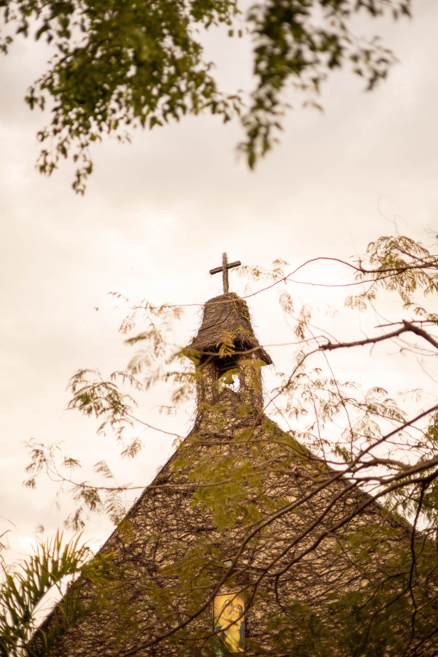 Moss-covered stone church steeple with a cross on top, framed by green tree branches against a cloudy sky.