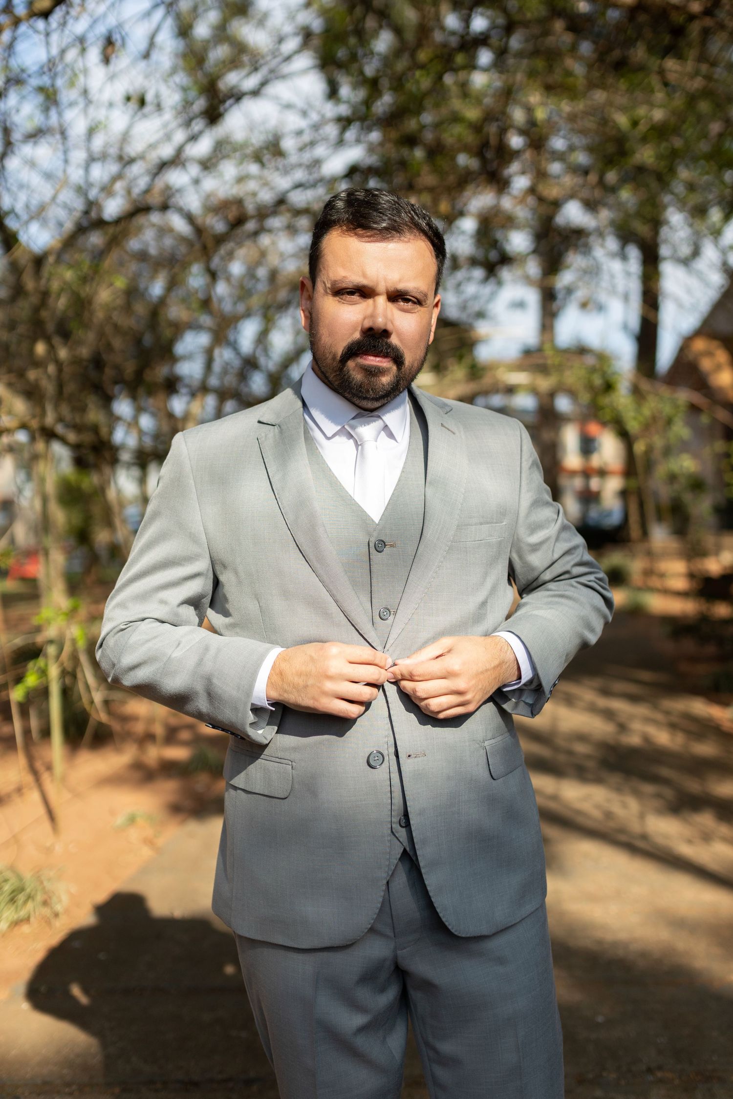 A bearded man in a gray three-piece suit buttoning his jacket outdoors among trees.