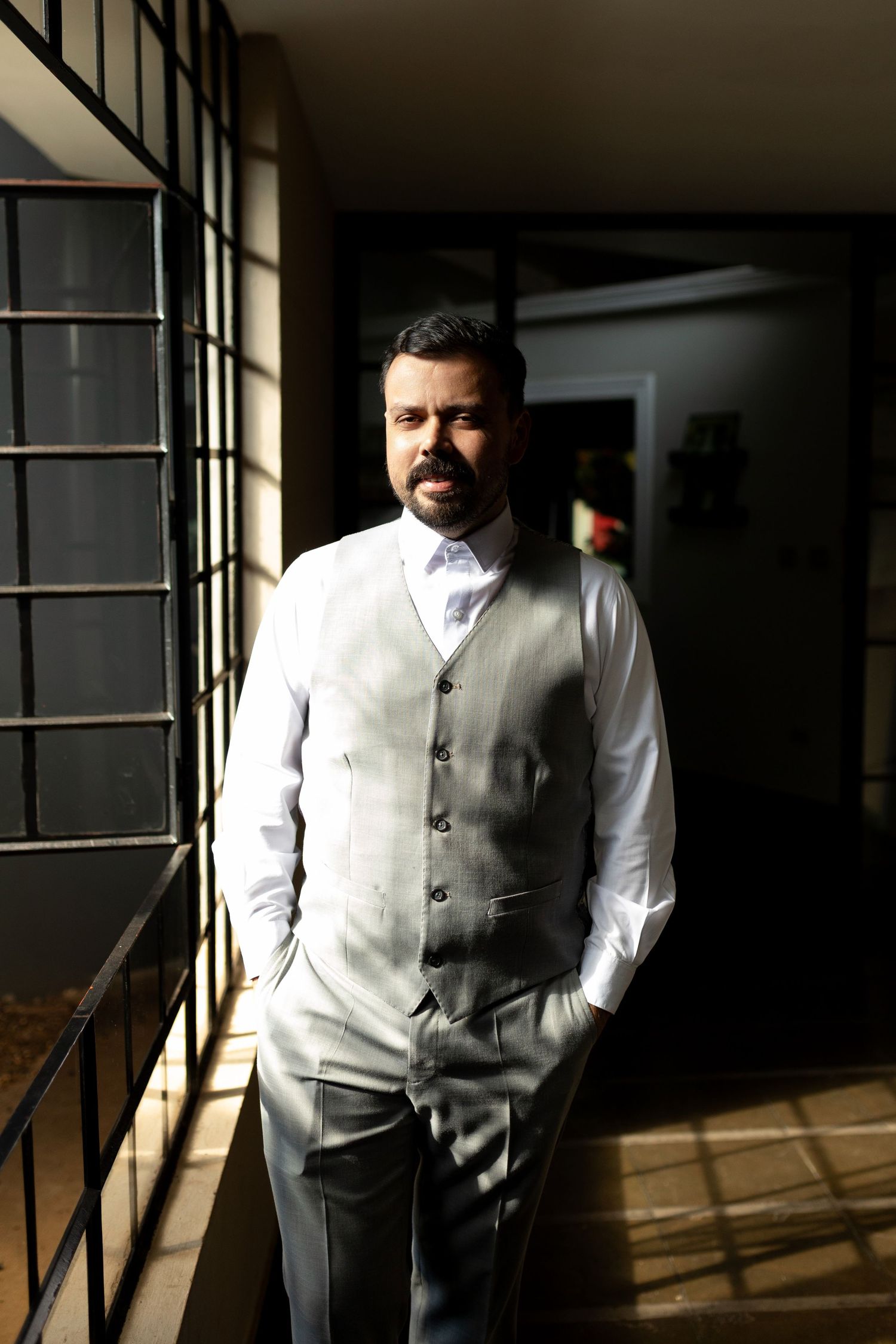 Man in gray vest and white shirt with bow tie stands confidently in sunlit hallway with hands in pockets.