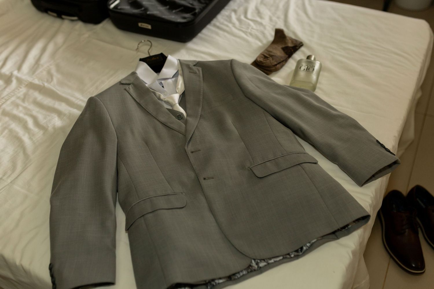 Gray men's suit jacket laid flat on a bed with dress shoes, white shirt, and luggage in the background.